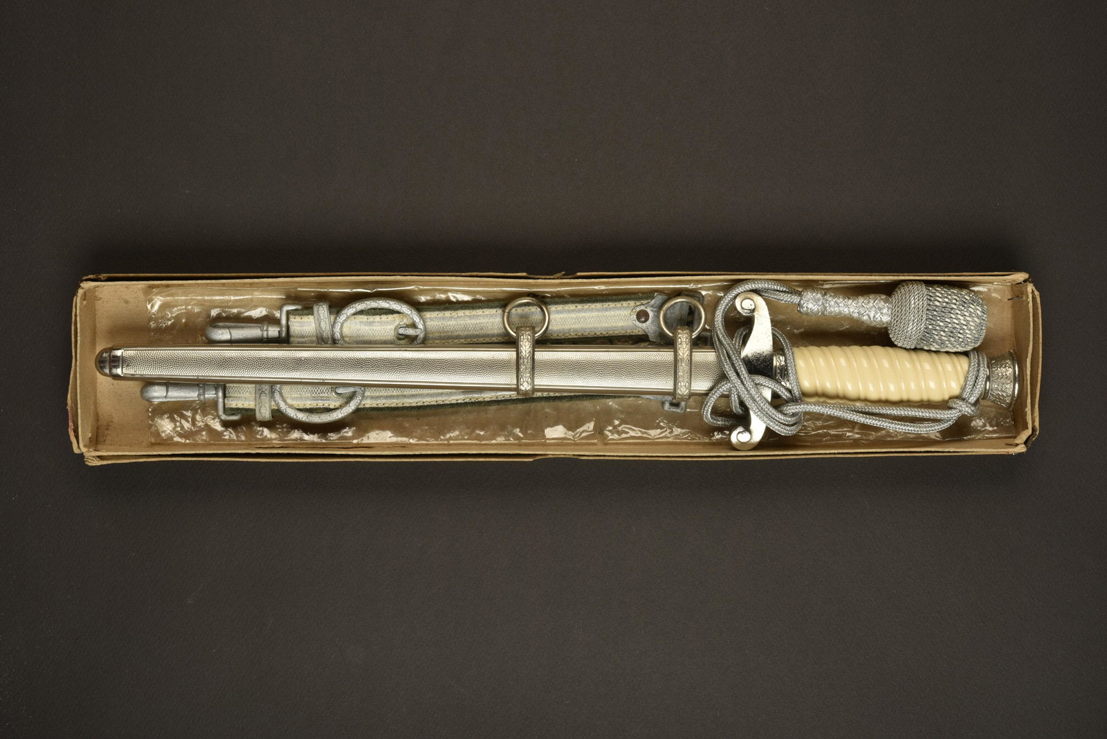 Dague d officier de la Heer en boite d origine. Heer: Garnitures en metal, couleur argent. Fusee de couleur blanche. Dragonne en fil d argent monte de maniere reglementaire. Fourreau en metal grenele, presentant une legere usure, bagues decorees de feuil