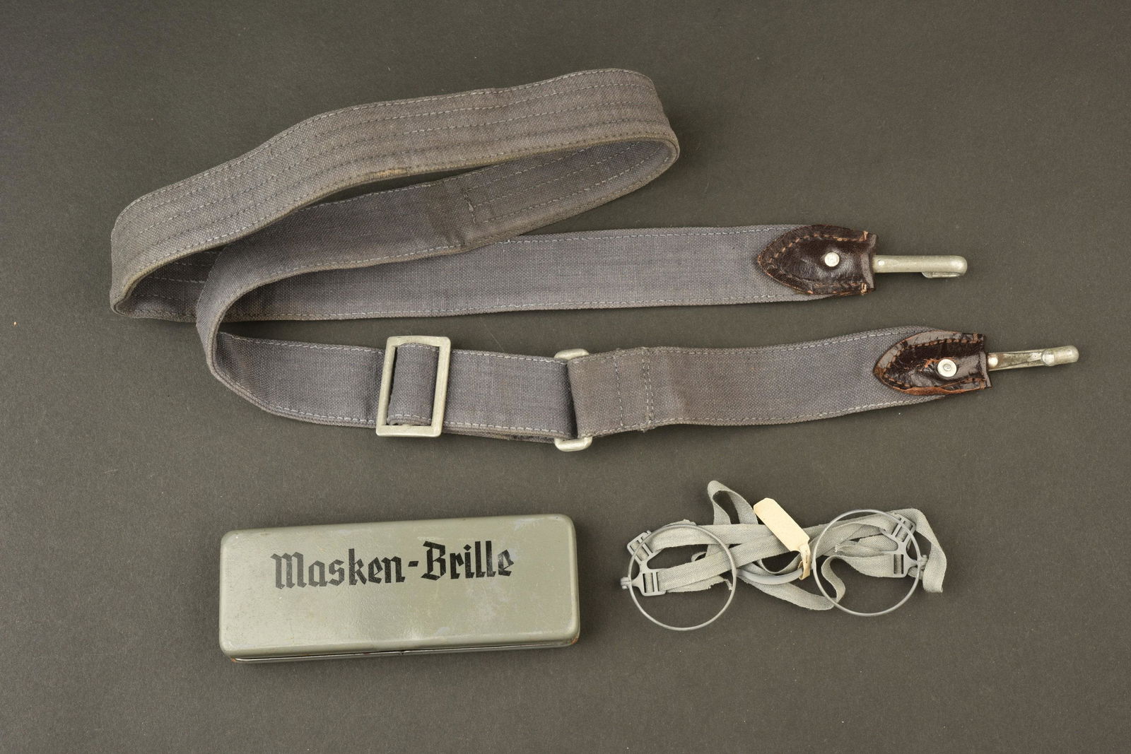 Ensemble d equipement allemand. German equipment set.: Comprenant une sangle de sac a pain en tissu gris bleu, un mousqueton est casse. Marquages LSAB 38. Une paire de lunette Masken Brille, boite en metal, marquages M6842. Lunettes completes, les verres