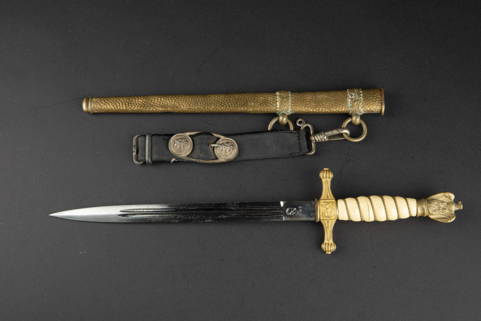Dague KM. Kriegsmarine dagger: Dague d officier Kriegsmarine. Garnitures en metal dore. Fusee de couleur blanche presentant quelques taches, filigrane present, nombreuses fissures. Dragonne absente. Fourreau martele, bagues decoree