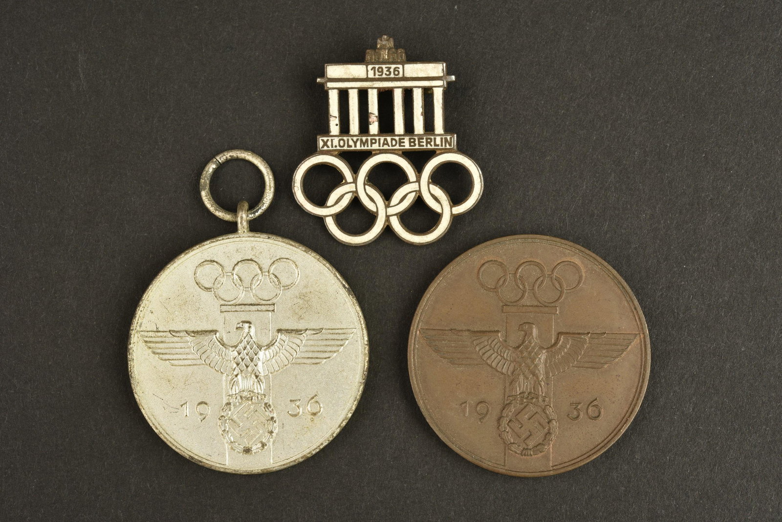 Insignes JO 1936. JO 1936 insignia: Insignes JO 1936. Comprenant un insigne en metal emaille blanc, Berlin 1936, attache complete, marquages Ges Gesch. Une medaille Fur Verdienstvolle Mitarbeit bei den Olympischen Spielen 1936, anneau p