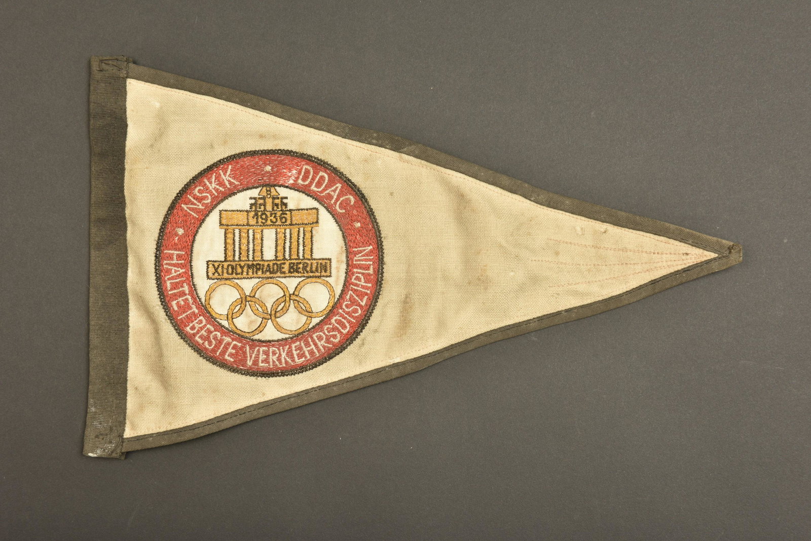 Fanion NSKK JO 1936. Pennant NSKK JO 1936.: Fanion NSKK JO 1936. En tissu, bordure en tissu noir, fond blanc. Insignes brode, marque NSKK DDAC Haltetbeste Verkehrsdisziplin. Biface. A noter une certaine usure et patine de la piece, ainsi que qu