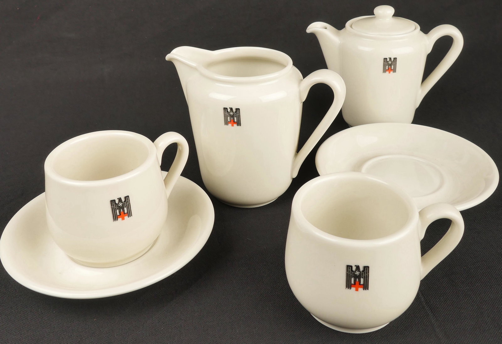 Vaisselle DRK. German DRK tableware: Vaisselle DRK. Comprenant un pot a lait en porcelaine blanche, anse et bec verseur complet, bel insigne de la DRK, logo fabricant present. Hauteur 12. Deux sous tasses en porcelaine blanche, sans marq