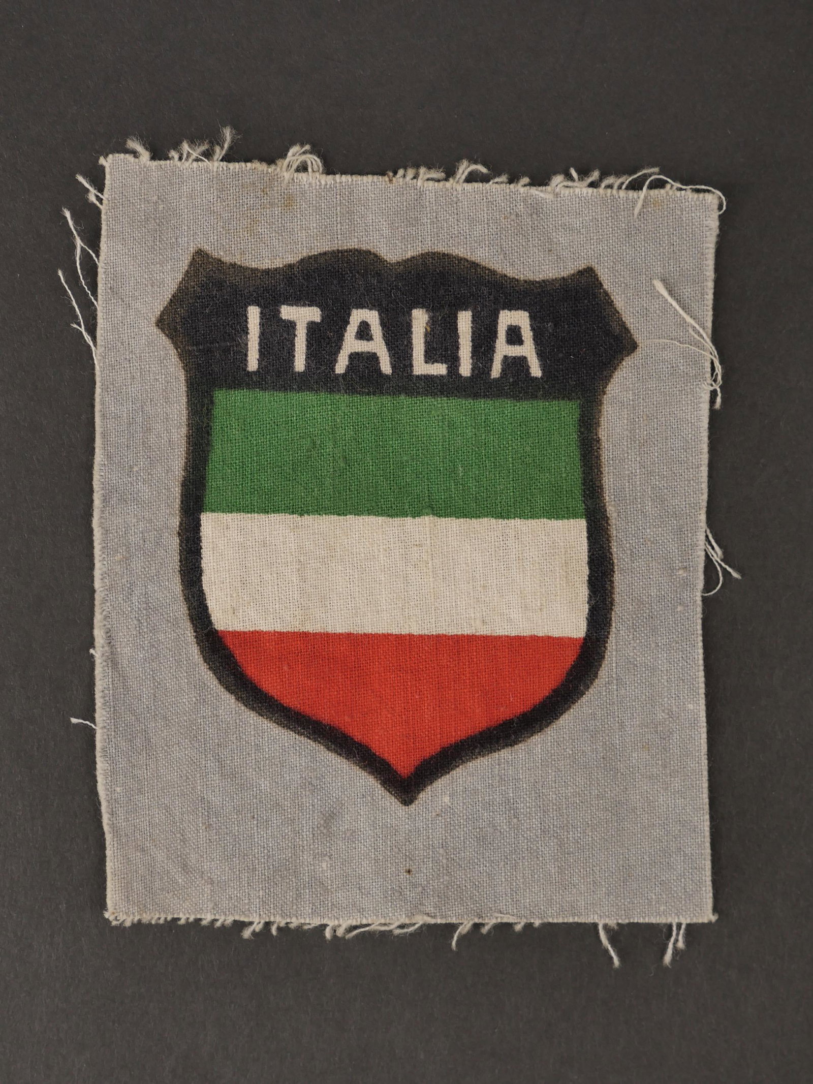 Insigne de manche italien. Italian sleeve badge.: Insigne de manche des volontaires italien dans la Heer. Modele imprime sur fond gris. Drapeau italien en forme d ecu surmonte de la mention ITALIA. Jamais monte, partiellement effiloche sur les bordur