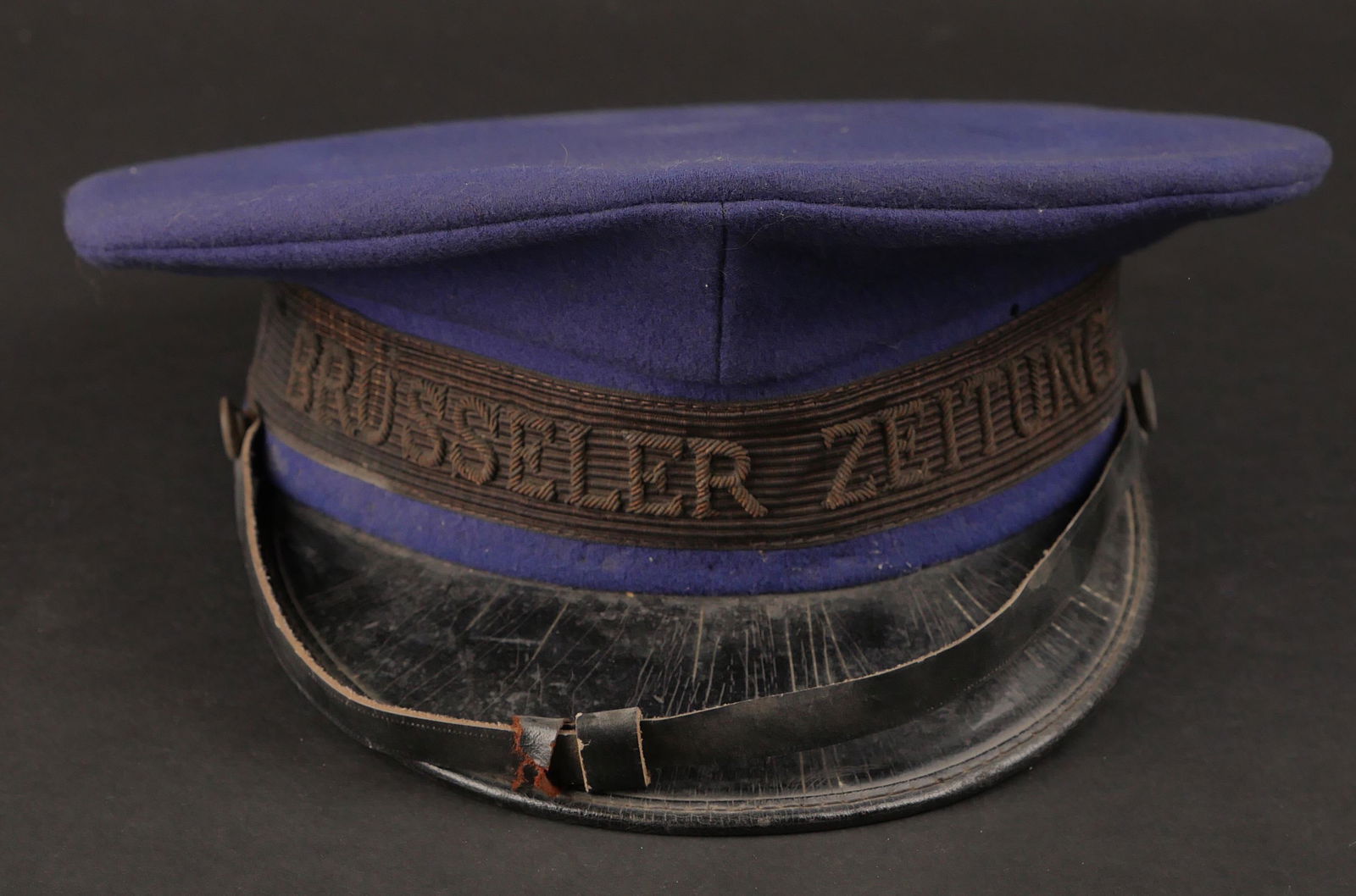 Casquette Brusseler Zeitung. Casquette Brussels Newspaper.: Casquette Brusseler Zeitung. En drap bleu marine. Bandeau en tissu noir, broderie en relief, Brusseler Zeitung. Fause jugulaire en cuir presente. Interieur double en tissu marron. Mica complet. Bandea
