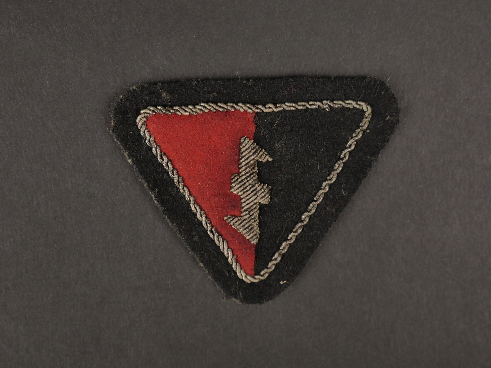 Insigne de coiffure NSB. NSB headgear badge: Insigne de coiffure du NSB. Modele en drap rouge et noir, monte sur fond noir. Insigne du NSB en cannetille argent, ainsi que le galon faisant le tour de l insigne. Dos de l insigne double d un papier