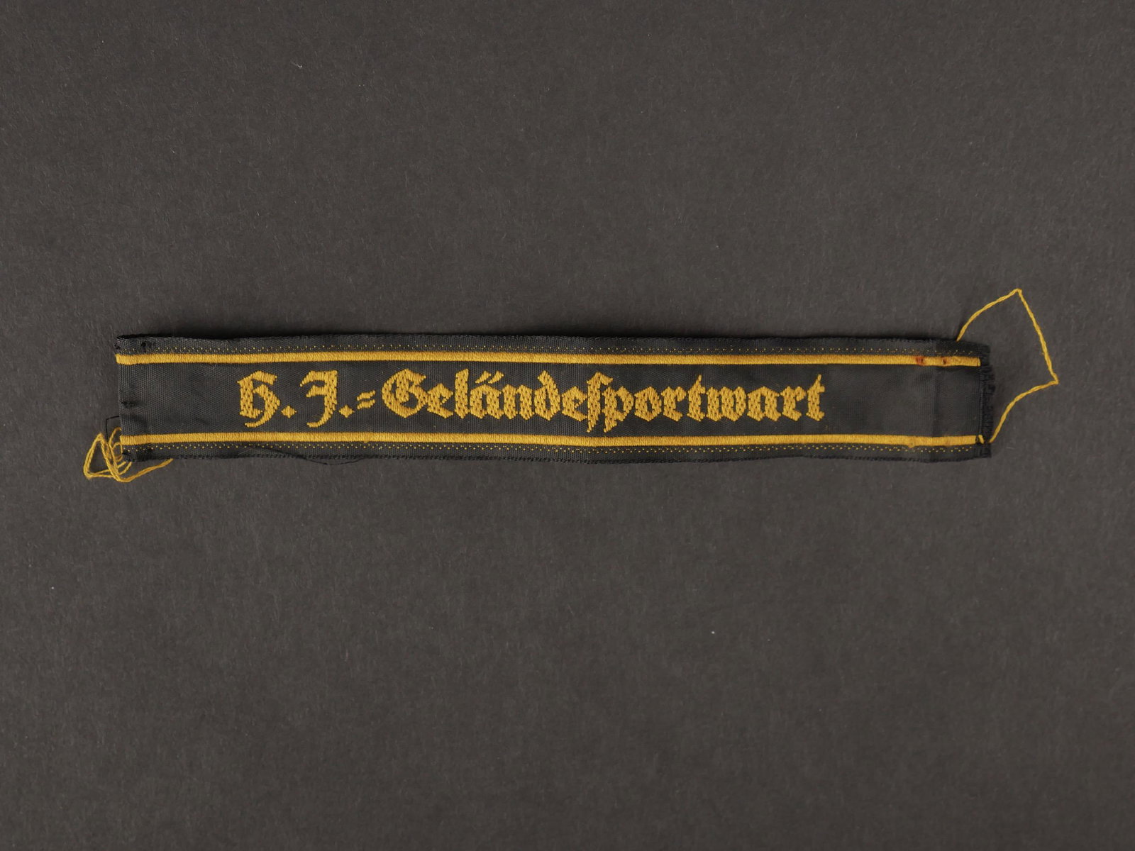 HJ Gelandesportwart.: Hitlerjugend Gelandesportwart. En tissu noir, tissage en fil jaune, marque HJ Gelansportwart. Longueur 14,5 cm. Jamais monte. A noter une legere usure et patine de la piece. Etat I . Hitlerjugend Gela