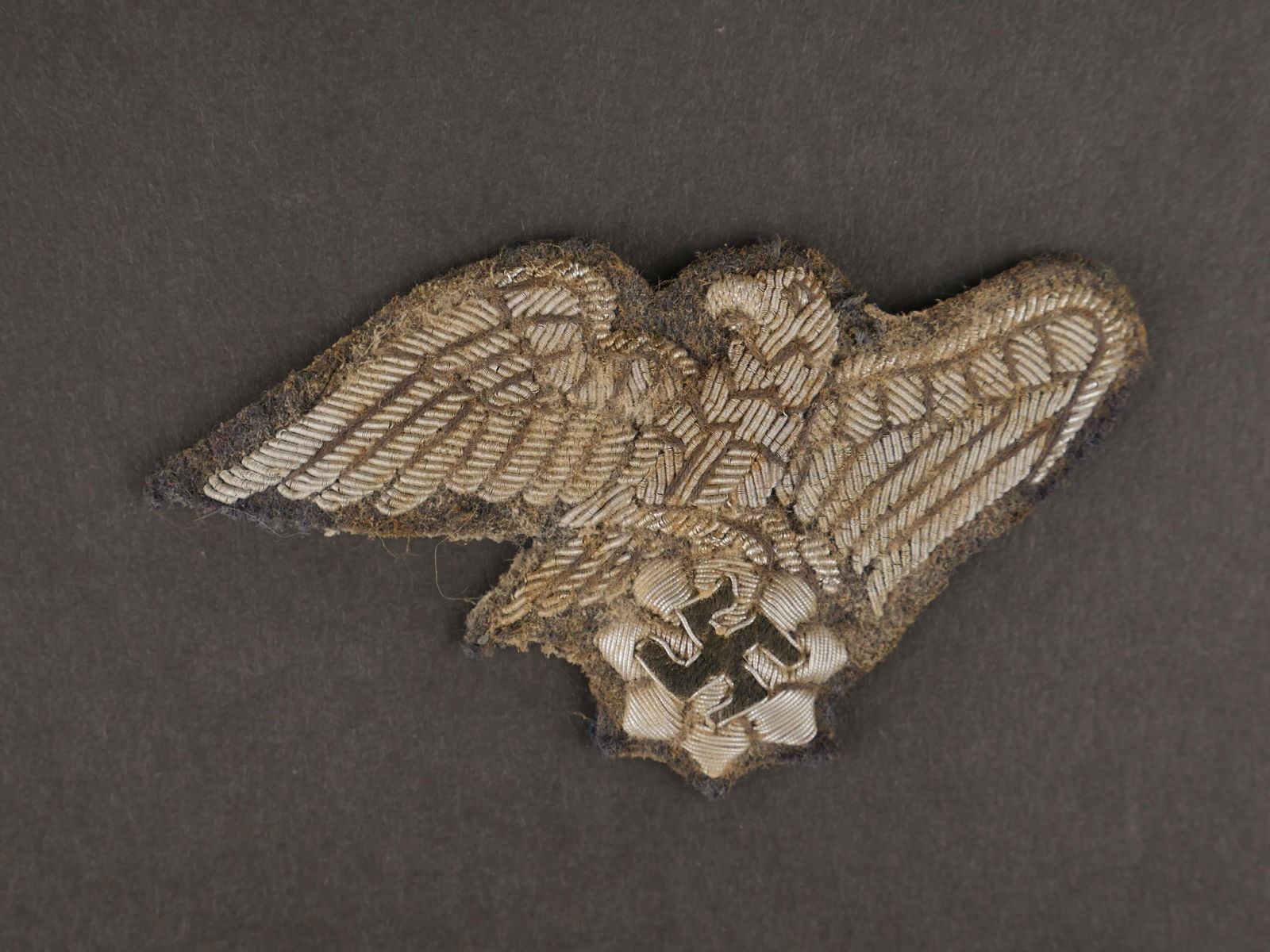 Aigle RLB. RLB Eagle.: Aigle de casquette RLB. En cannetille argent sur fond gris. Dos de la piece renforce par un papier noir. A noter une certaine usure et patine de la piece. Etat II+. Eagle of cap RLB. In silver canneti