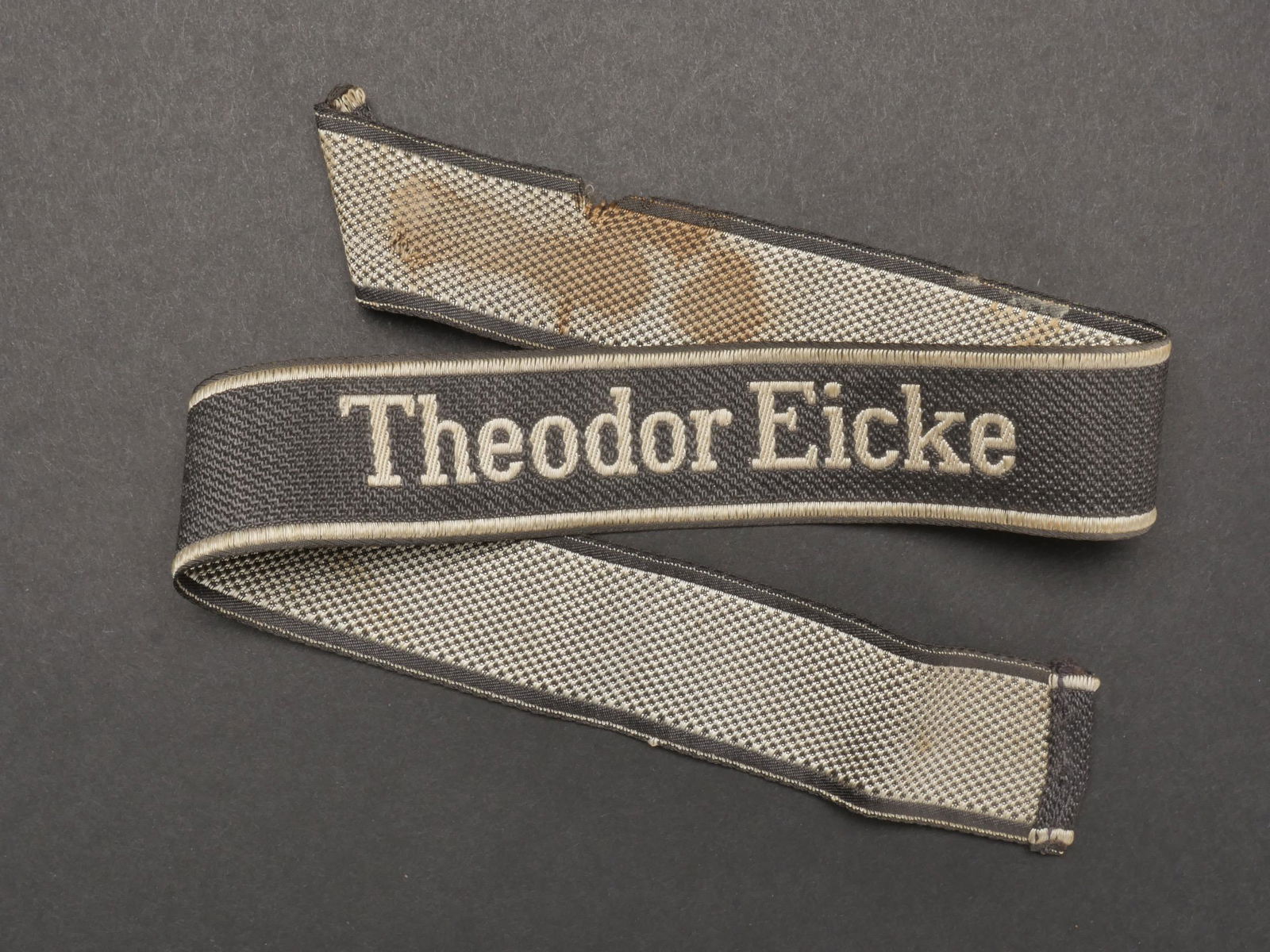 Bande de bras Theodor Eicke. Theodor Eicke cuff tittle.: Bande de bras Theodor Eicke. Bande en tissu noir. Broderie en fils blanc Theodor Eicke. Galon tisses en fils blanc de chaque cote de la bande de bras. Marquages Bevo Wupperthal. Quelques taches et un