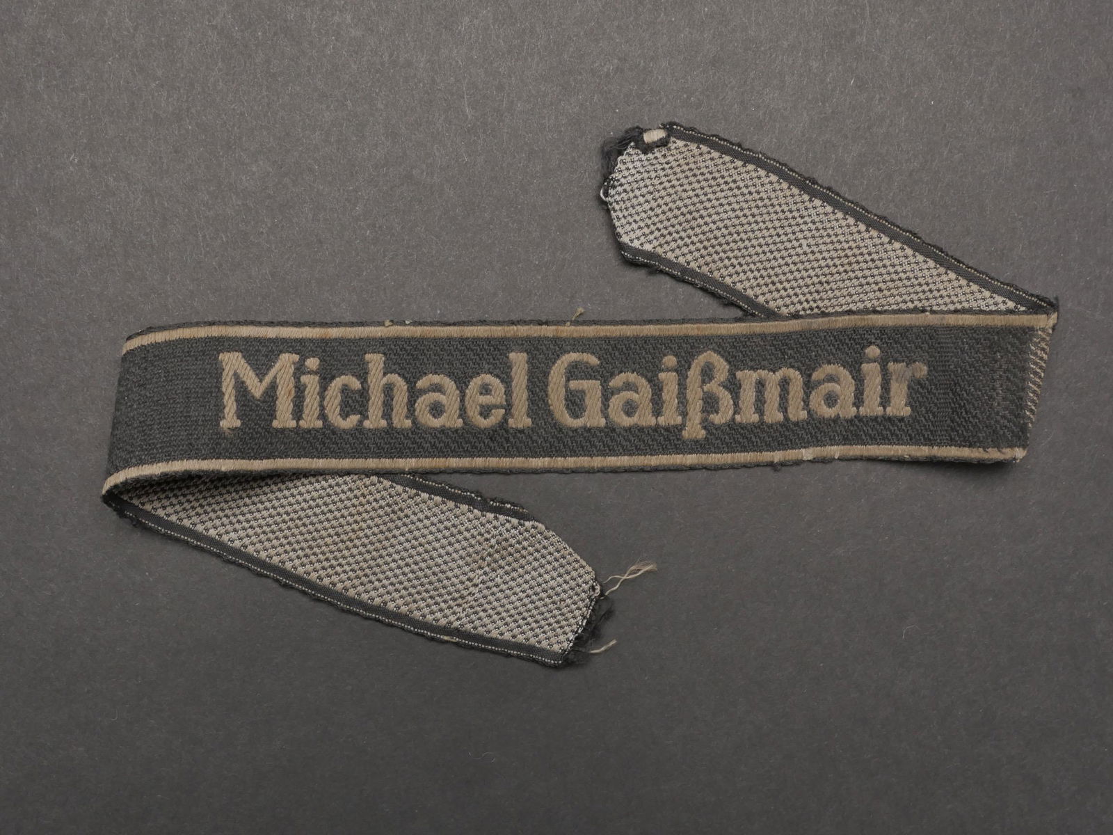 Bande de bras Michael Gaissmair. Michael Gaissmair cuff tittle.: Bande de bras Michael Gaissmair. Bande en tissu noir. Broderie en fils blanc Michael Gaissmair. Galon tisses en fils blanc de chaque cote de la bande de bras. Incomplete. Nombreuses marques d usures e