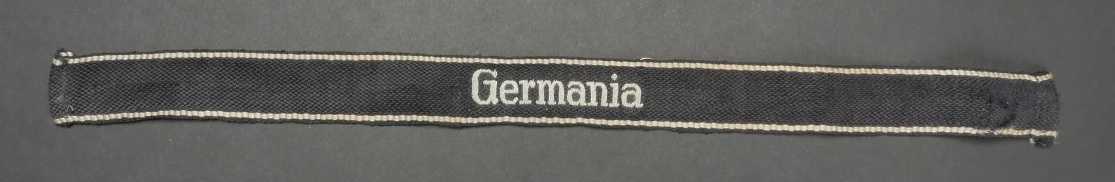 Bande de bras Germania. Germania cuff tittle.: Bande de bras Germania. Modele tisse, couleur argent. Marquages Germania, renfort en tissu noir au dos du marquage. Deux lisere argent tisses. Longueur 46 cm. A noter une certaine usure et patine de l