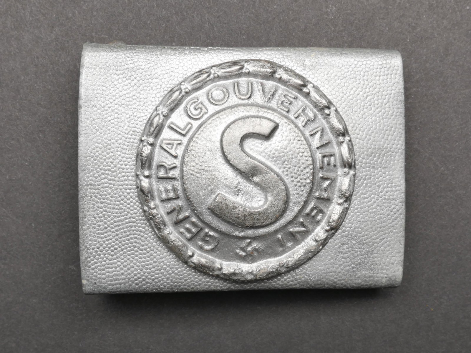 Boucle de ceinturon General Gouvernement. General Gouvernment belt buckle.: Boucle de ceinturon General Gouvernement. En metal grenele. Fabrication en deux parties. Legerement deformee. Boucle a deux ardillons complete. Sans marquages fabricant visibles. A noter une certaine 