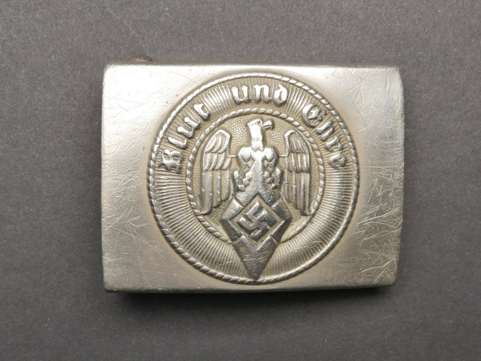 Boucle de ceinturon HJ. HJ belt buckle.: Boucle de ceinturon Hitlerjugend. En metal, anodise. Trace de colle au dos. Insigne et devise en relief. Trace de colle au dos. Boucle a deux ardillons incomplete. Fabrication RZM 17 Assmann Ges Gesch