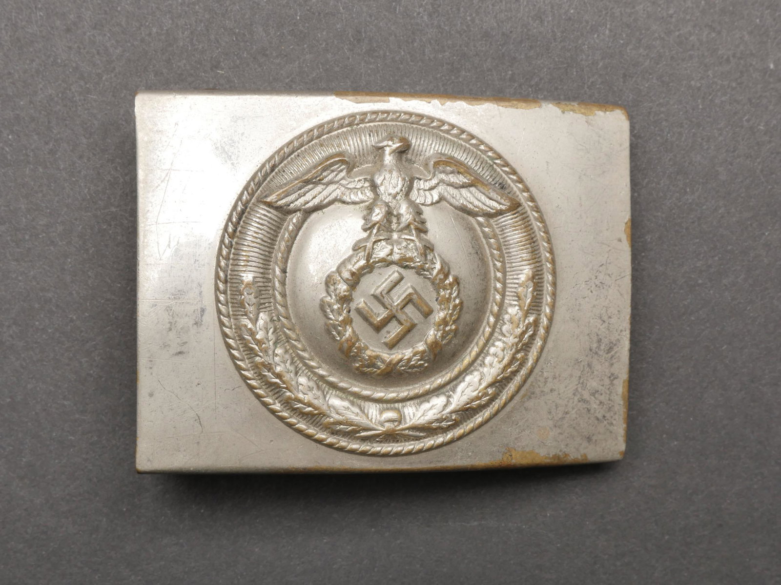 Boucle de ceinturon NSKK. NSKK belt buckle.: Boucle de ceinturon NSKK. En metal nickele. Insigne en relief, presentant des marques d usures. Boucle a deux ardillons complete. Sans marquages visibles. A noter une certaine usure et patine de la pi