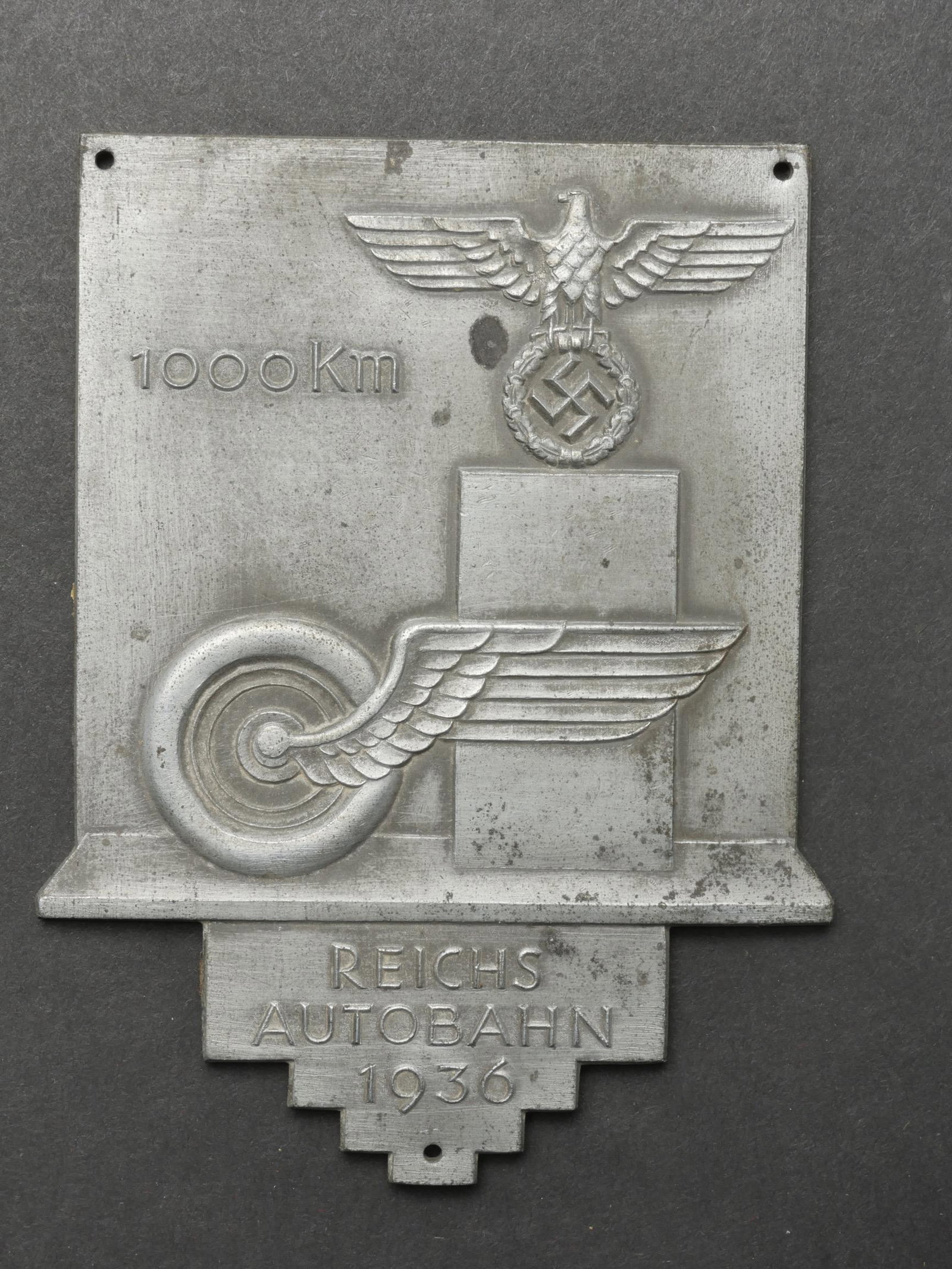Plaquette Reichsautobahn 1936. Reichsautobahn plate.: Plaquette Reichsautobahn 1936. En metal couleur argent a 70%. Decors en relief, 1000 km Reichsautobahn 1936. Trois points de fixation. Traces de colle au dos. Dimensions 11 x 7,5 cm. A noter une certa