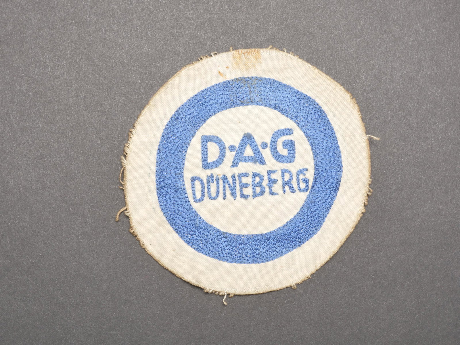 Insigne DAG. DAG insignia.: Insigne DAG. Modele tisse et brode sur fond blanc. Brode DAG Duneberg. A noter une certaine usure et patine de la piece, ainsi que des taches. Diametre 11 cm. Etat II+. DAG badge. Woven and embroidere