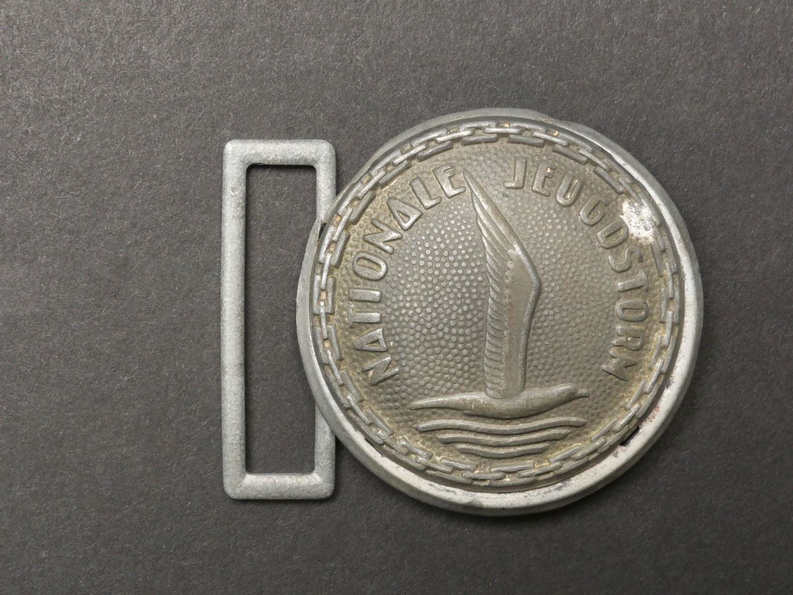 Boucle de ceinturon Jeugdstorm. Jeugdstorm belt buckle.: Boucle de ceinturon Jeugdstorm. En metal, fabrication en deux parties. Un passant present. Boucle a deux ardillon manquante. A noter une certaine usure et patine de la piece. Etat II+. Jeugdstorm belt