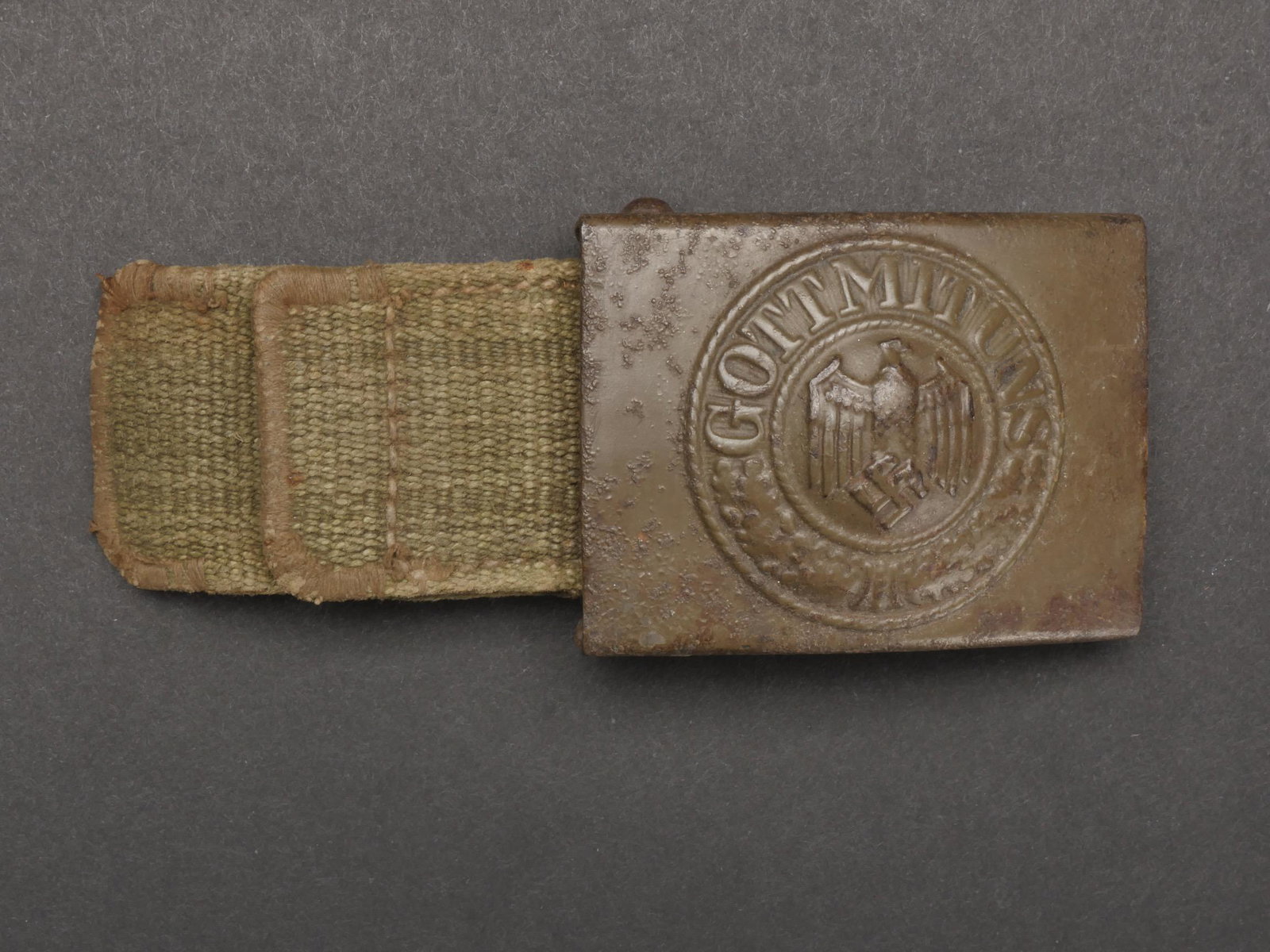 Boucle de ceinturon AK. Belt buckle AK.: Boucle de ceinturon AK. En metal. Peinture Feldgrau a 60%. Fortement oxydee. Sans marquages visibles. Patte en toile Feldgrau, sans marquages visibles. A noter une certaine usure et patine de la piece
