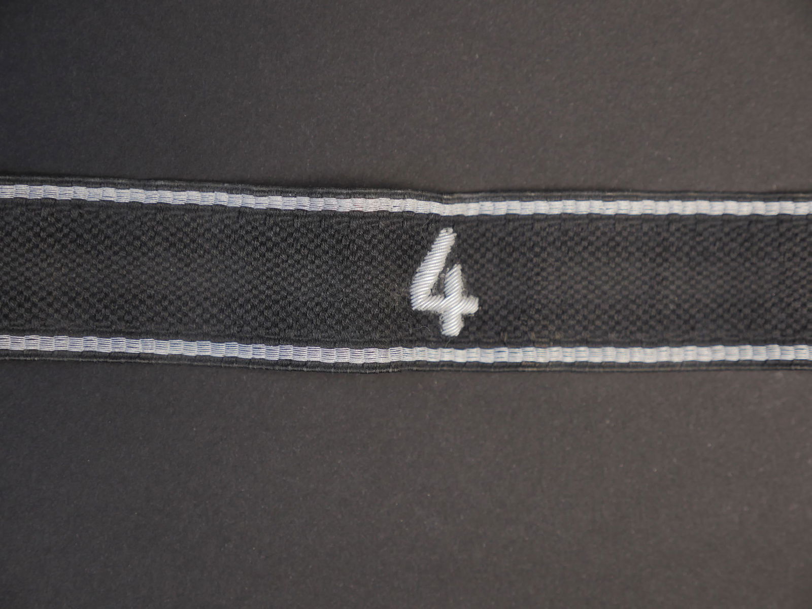 Bande de bras SS. SS cufftittle.: Bande de bras SS. En tissu noir, deux liseres argent. Chiffre 4 brode en cannetille argent. Longueur 46,5 cm. A noter une certaine usure et patine de la piece. Etat II+.SS arm band. Black fabric, two