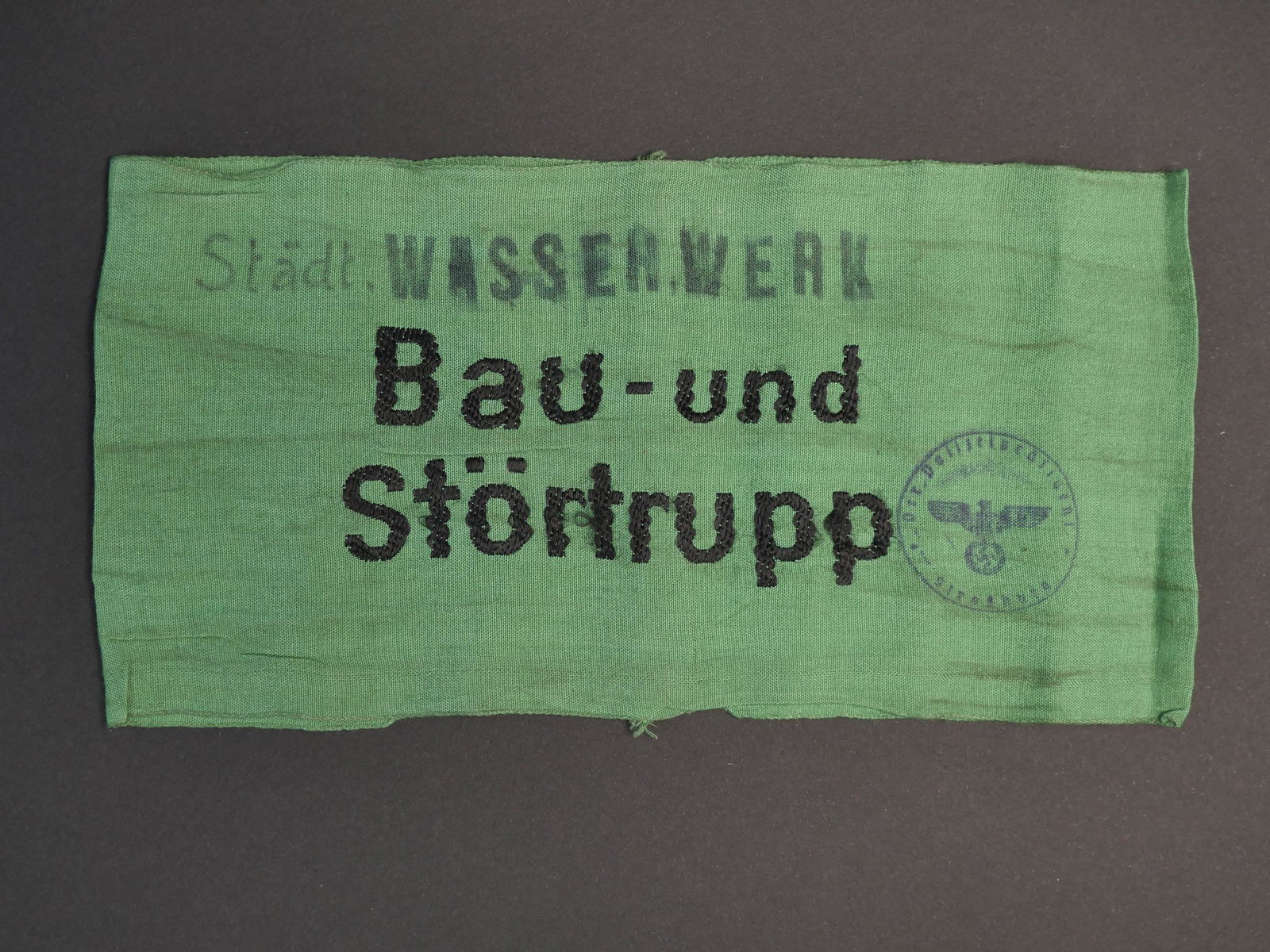 Brassard Bau. Bau armband.: Brassard Bau und Stortrupp. En tissu coton vert. Broderie noire Bau und Stortrupp. Inscription manuelle et au tampon St adt Wasser Werk, tampon allemand de la ville de Strasbourg. A noter une certaine