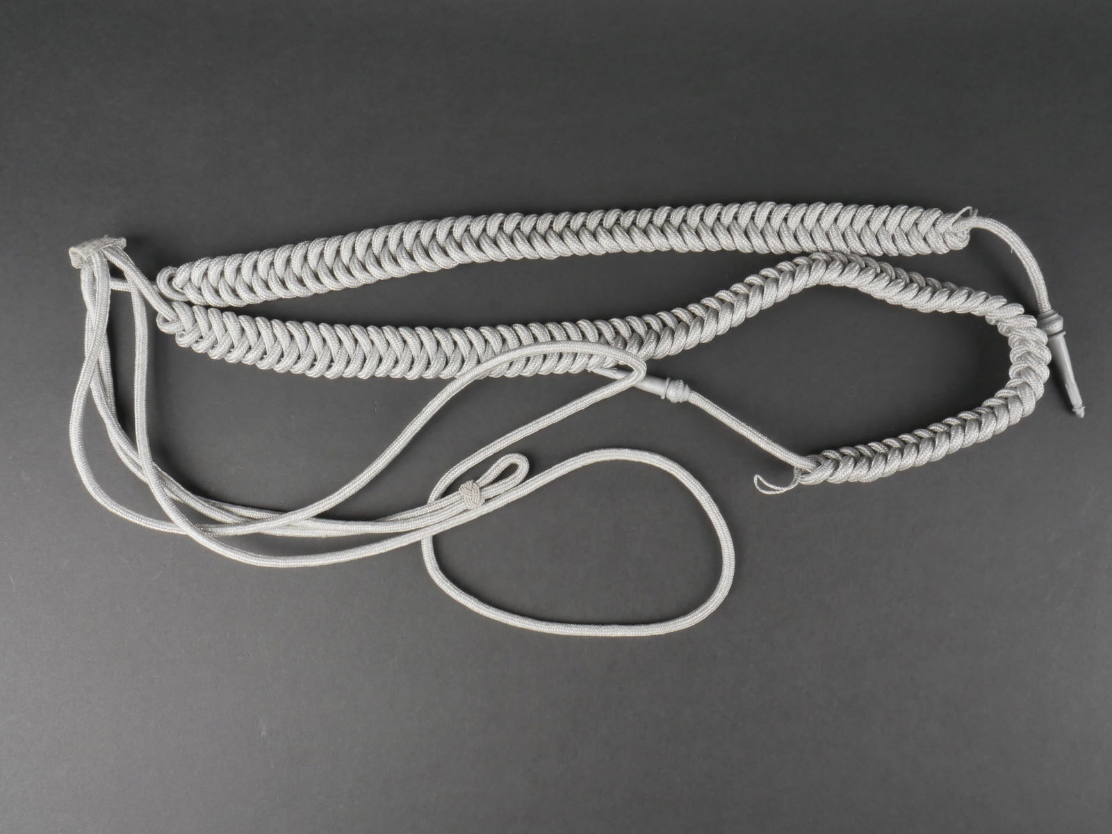 Aiguillette Heer. Heer aiguillette.: Aiguillette de la Heer. En fils tresses argent. Les attaches aux boutons sont presentes. Les glands sont en metal, egalement couleur argent. A noter une certaine usure et patine de la piece. Etat II+.