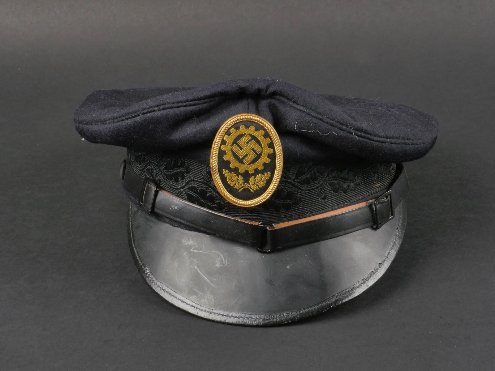 casquette du DAF. German DAF cap.: casquette du DAF. coiffe en drap noir. Bandeau decore de feuilles de chene. Visiere en tres bon etat. Fausse jugulaire en cuir maintenue par deux boutons recouverts de tissu noir. Insigne du DAF tisse