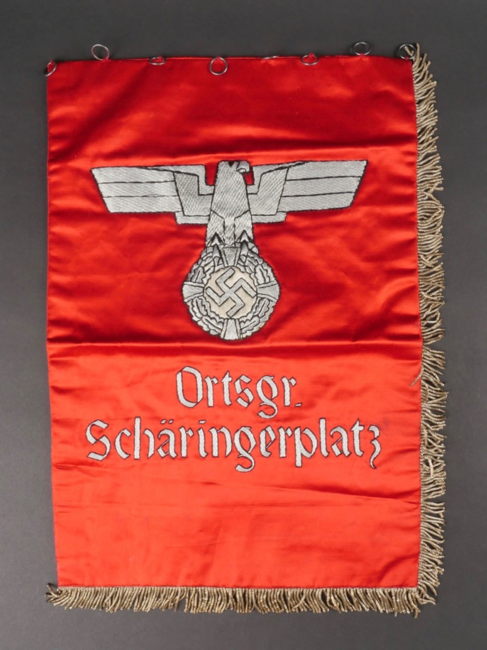 Drapeau Orstgruppe Scharingerplatz. Orstgruppe Scharingerplatz flag.: Drapeau Orstgruppe Scharingerplatz. En epais tissu soyeux rouge. Frange sur deux cotes. Les anneaux de fixation sont presents. Bel aigle allemand en epaisse broderie couleur argent. Marquage egalement