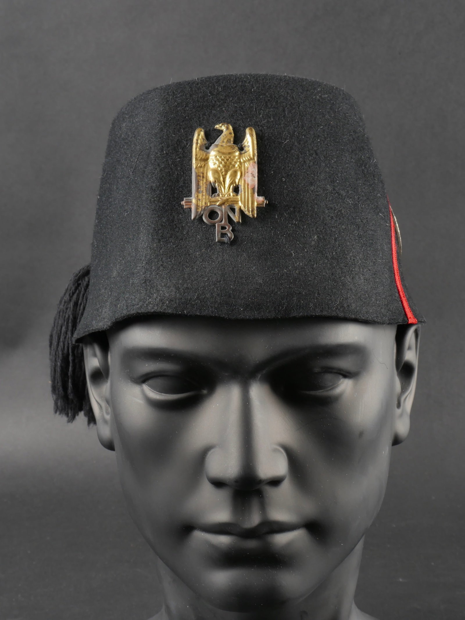 Fez de Ballila Scelto. Fez by Ballila Scelto.: Fez de Ballila Scelto. En feutre noir avec son insigne metallique frontal, pompon, losange de grade au milieu duquel est cousu un medaillon sportif au profil du Duce des es Opera Balilla. Traces d’u