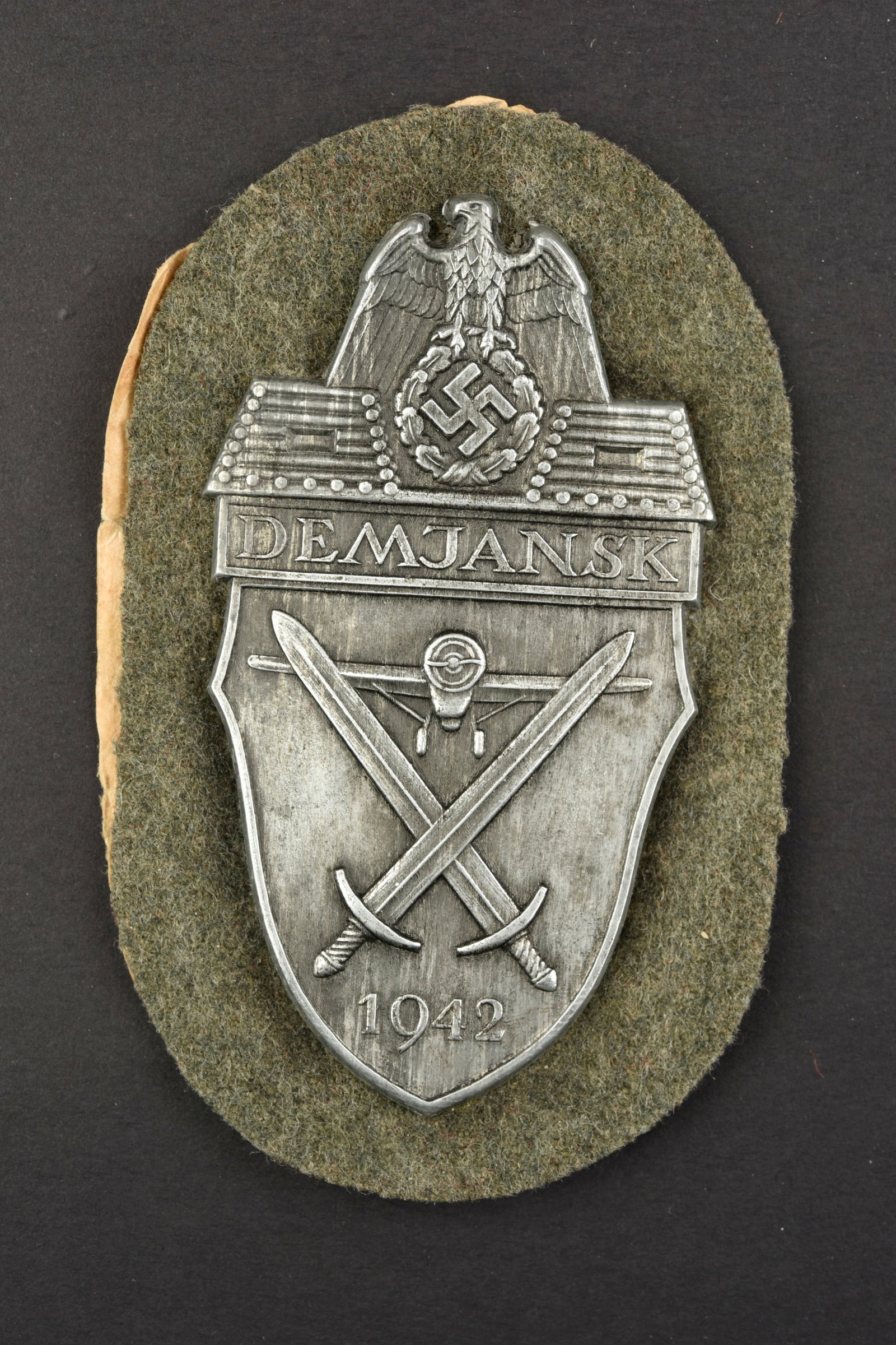 Plaque Demjansk. Demjansk insignia.: Plaque Demjansk. EN metal bien marquee Demjansk 1942, montage sur un support en drap Feldgrau. Contre plaque en metal complete. Papier au dos dessolidarise. A noter une certaine usure et patine de la 