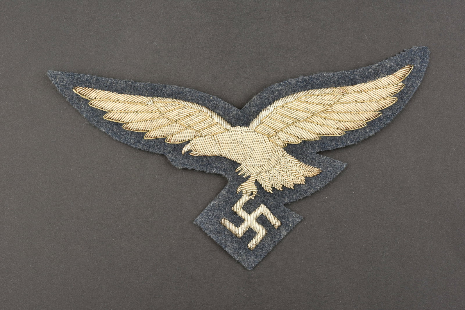 Aigle de cape LW. Eagle cape LW.: Aigle de cape de la Luftwaffe. En epaisse cannetille argent, sur fond en drap gris/bleu. Dos de la piece double de papier noir. Longueur 19 cm. A noter une certaine usure et patine de la piece, ainsi 