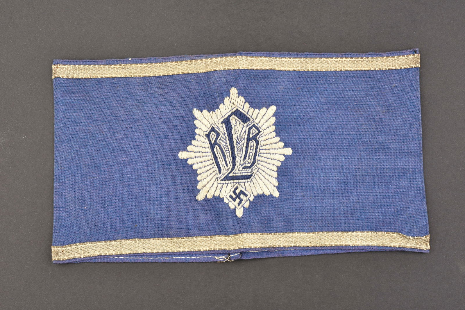 Brassard RLB. RLB armband.: Brassard RLB. Premier modele. En tissu coton bleu ciel. Insigne RLB tisse. Marquages au dos Ges Gesch. A noter une certaine usure et patine de la piece, ainsi que quelques taches. Etat II+. RLB cuff.