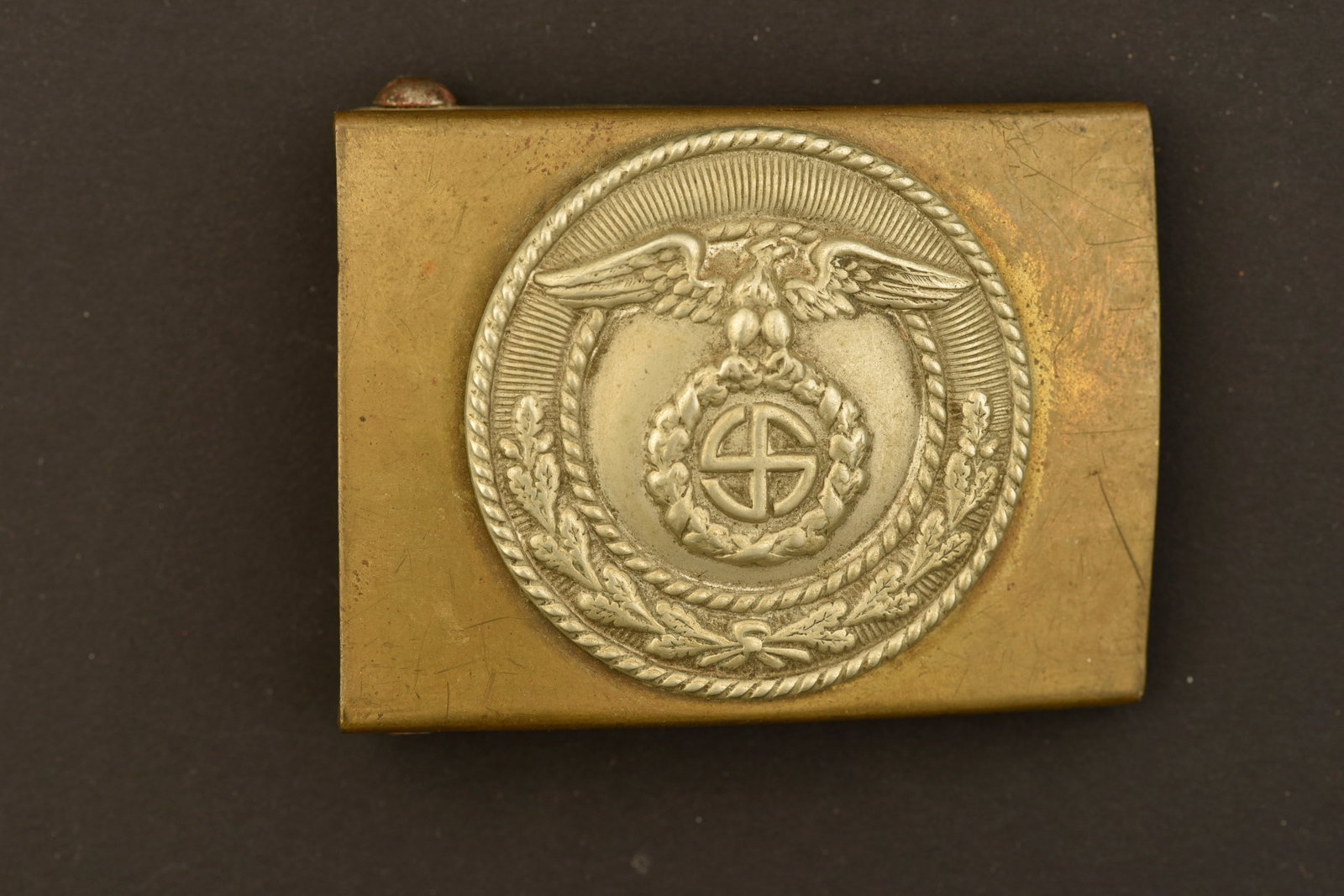Boucle de ceinturon SA. SA belt buckle.: Boucle de ceinturon SA. En metal laiton et metal. Fabrication en deux parties. Un bouchon en liege est colle au dos de la boucle. Toute la boucle est deformee. Insigne en relief. Boucle a deux ardillo