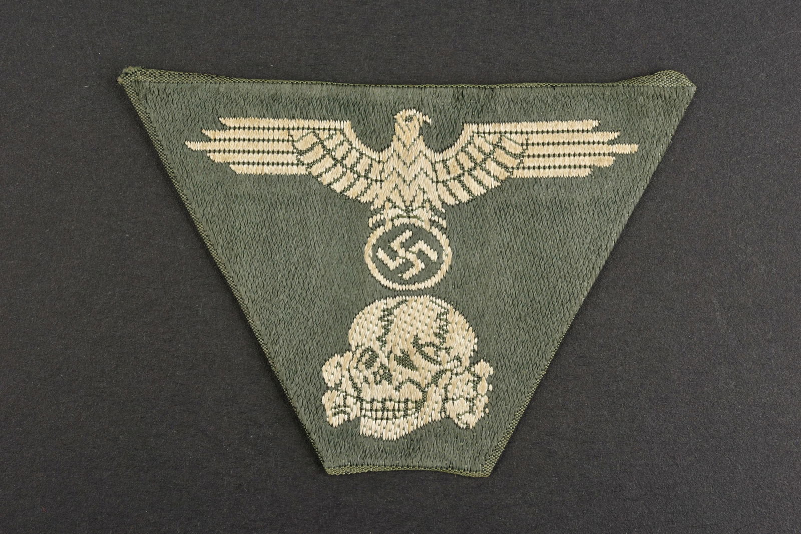 Insigne de coiffure SS. SS Cap insignia.: Insigne de coiffure SS. Insigne trapezoidal, representant une tete de mort et unaigle. Fond en tissu coton Feldgrau. Sans marquages visibles.a noter une certaine usure et patine de la piece. Etat II+.