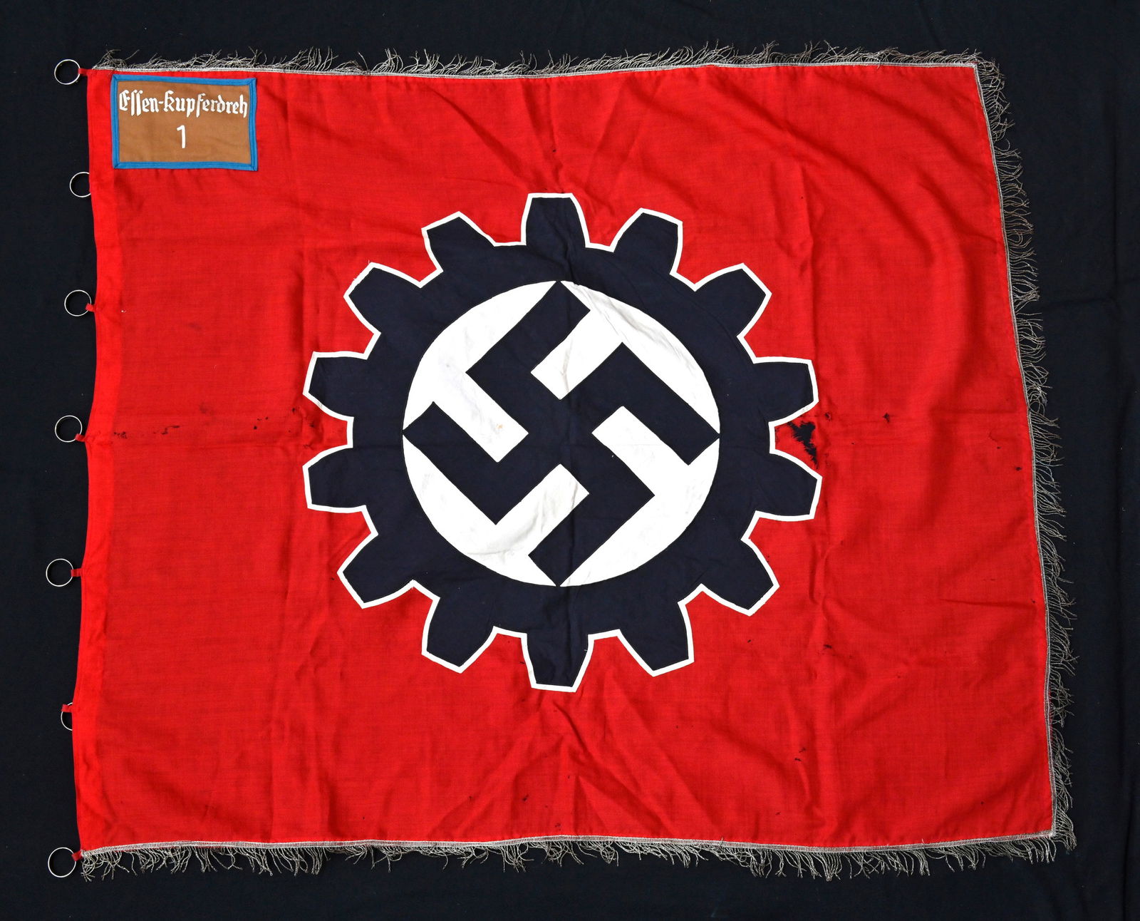 Drapeau du DAF. German DAF flag.: Drapeau du DAF. En tissu coton rouge. Insigne du DAF rapporte. Biface. Cartouche en drap marron, lisere bleu, marque Essen Kupferdreh 1. Frange sur trois cotes. Lesanneaux de fixationau mat sont prese
