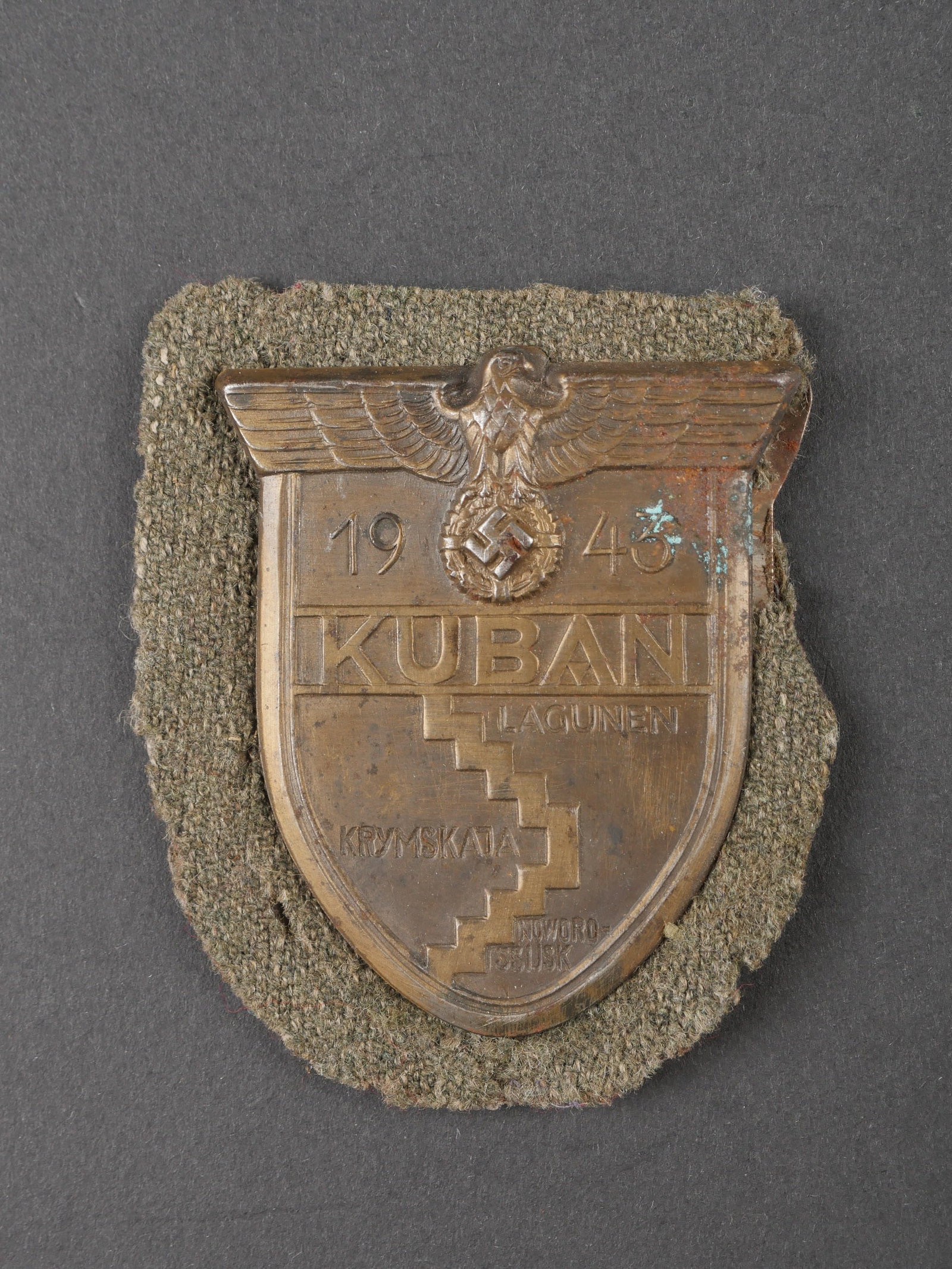 Plaque de bras Kuban. Kuban arm patch.: Plaque de bras Kuban. En metal embouti, bien marque Kuban 1943. Dos de la piece double en cuir noir. Feutrine Feldgrau complete. Contre plaque complete avec ses pattes de serrage. Feutrine fortement a