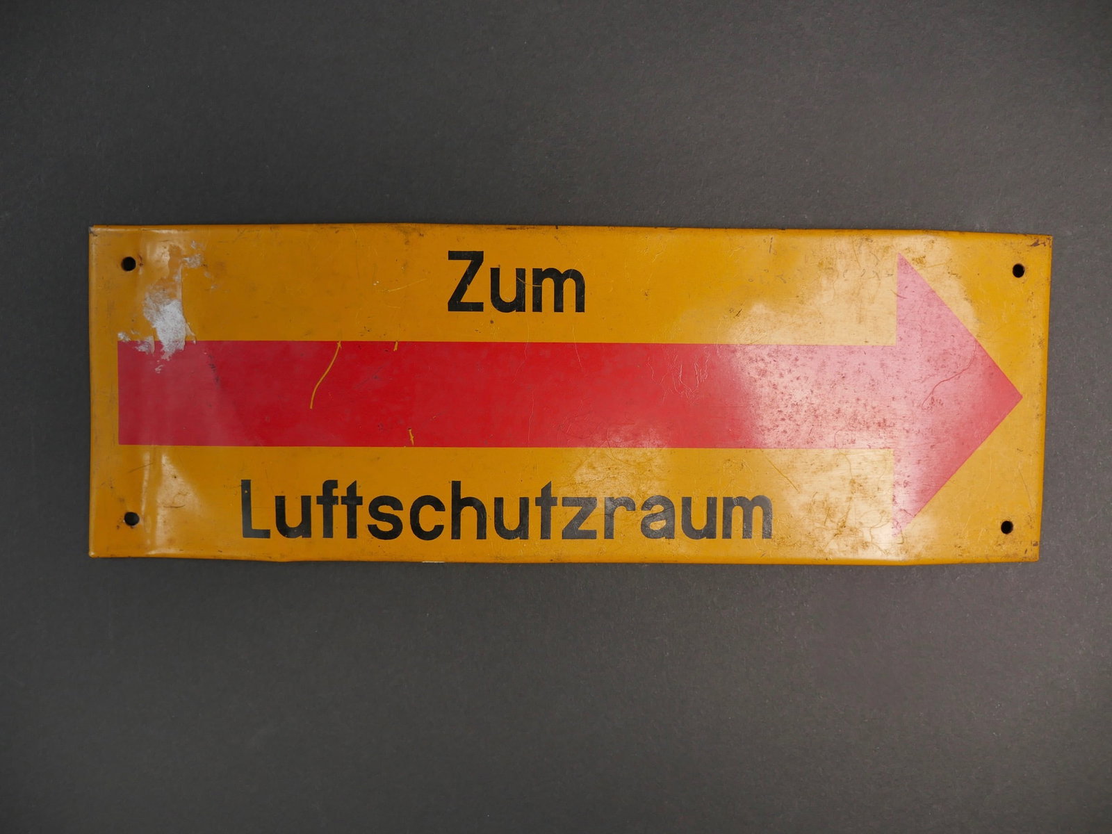 Plaque Luftschutz. Luftschutz plate.: Plaque indiquant un abri de la Luftschutz. En metal replie. Couleur jaune a 80%. Fleche rouge a 80%. Marquages Zum Luftschutzraum, en lettrage noir. Quatre points de fixation. Quelques bosses et defor