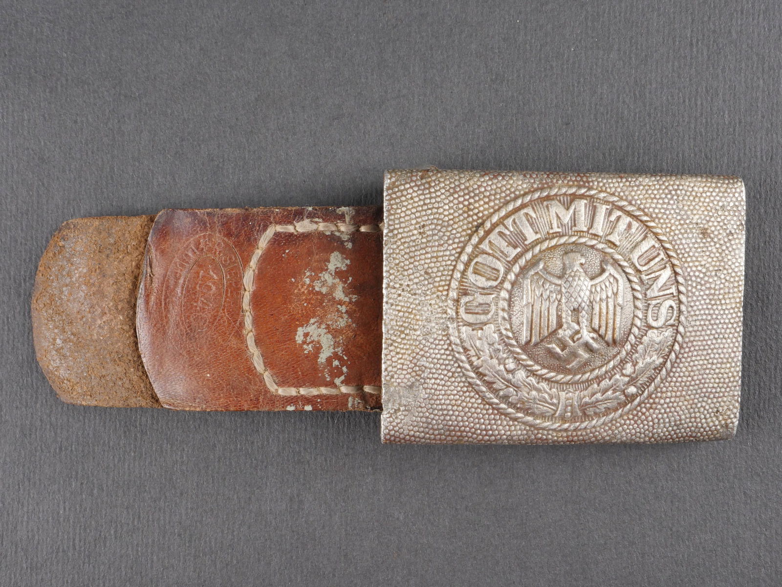 Boucle de ceinturon Heer. Heer belt buckle.: Boucle de ceinturon Heer. Modele en aluminium presentant de nombreuses bosses. Fabrication KQ, datee 1939. Patte en cuir cousue d’origine, datee 1940. Presente des traces d’usure et d’oxydation.