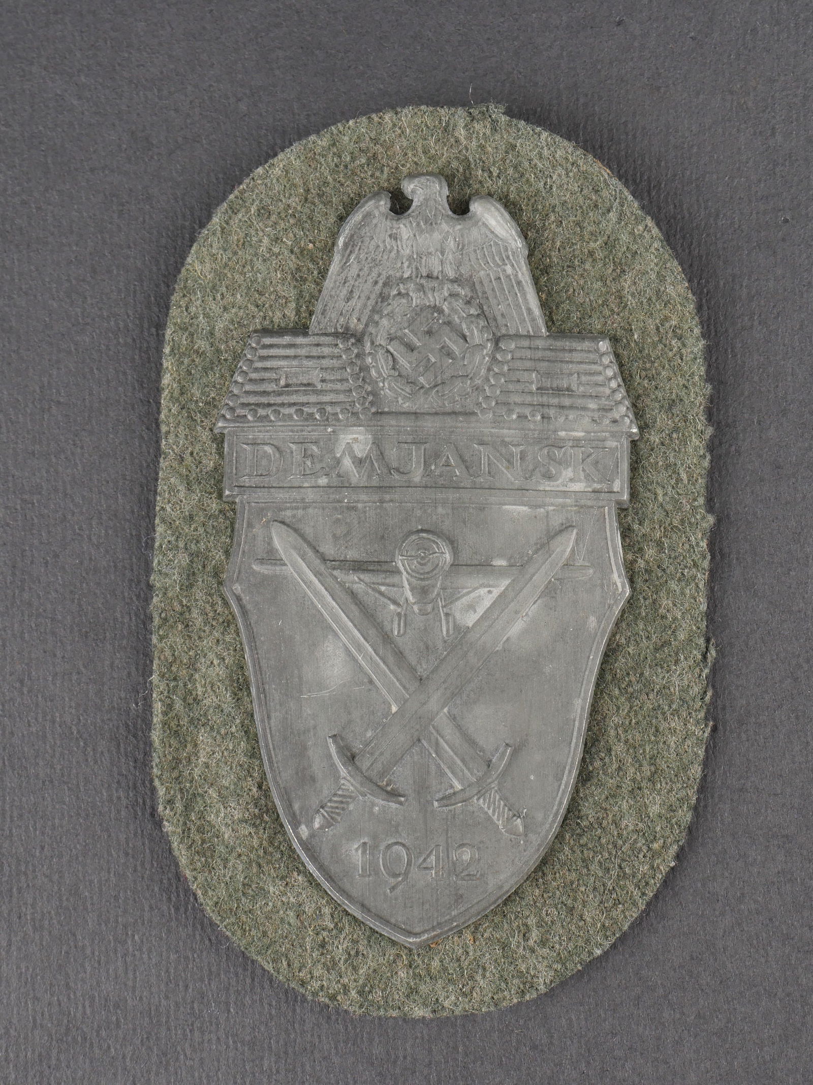 Plaque Demjansk. Demjansk insignia.: Plaque Demjansk en metal, a beaux decors en relief, fixee sur un support en drap feldgrau. Revers double d’un epais papier marron. Presente une certaine usure et patine. etat II+. Demjansk shield in