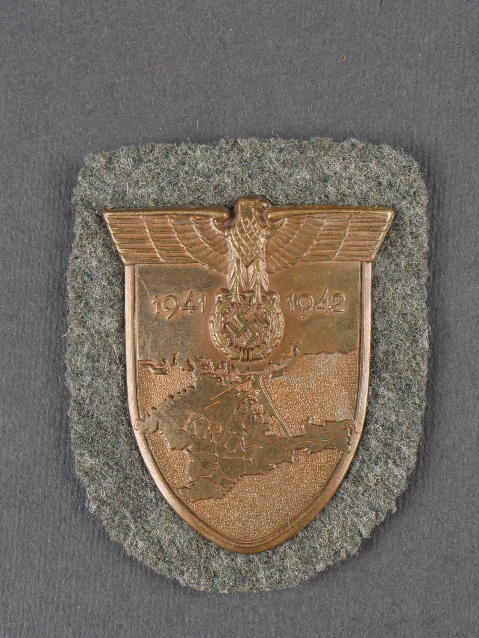 Plaque Krim. Krim shield: Plaque Krim en metal de couleur bronze, bien marquee Krim 1941 1942. Contre-plaque complete. Une attache est manquante. Drap feldgrau solidement fixe entre l’insigne et la contre-plaque. a noter une