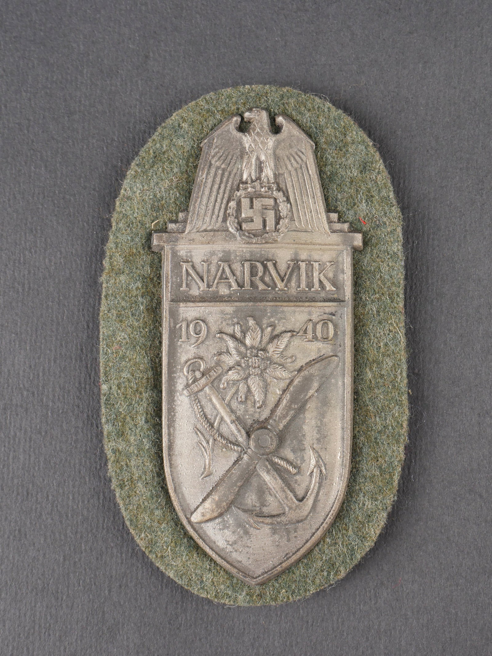 Insigne Narvik. Narvik shield.: Insigne de manche Narvik en metal a decor en relief, marque Narvik 1940. Support en drap feldgrau, contre-plaque metallique recouverte d’un second drap feldgrau colle. Presente une certaine usure et