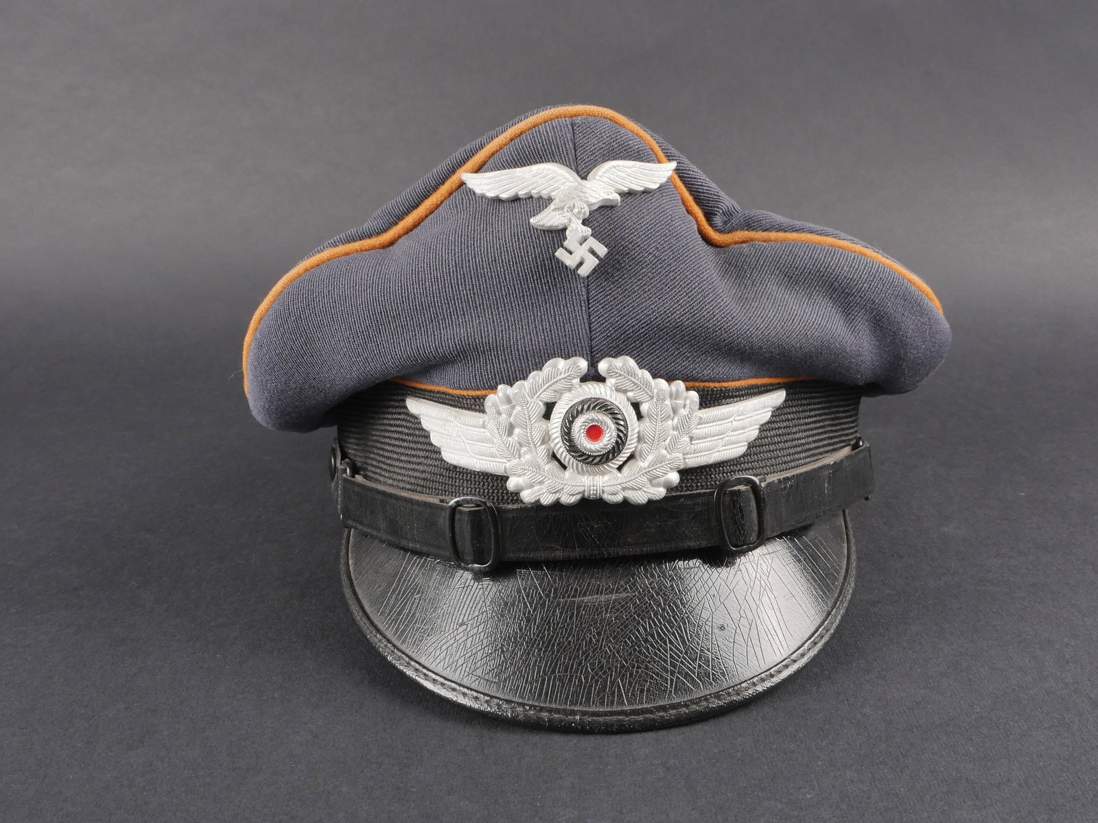 Casquette Luftwaffe. Luftwaffe visor cap.: Casquette de sous-officier des transmissions de la Luftwaffe, en gabardine bleu-gris, avec triple lisere marron. Bandeau en mohair noir, fausse jugulaire en cuir noir. Insignes metalliques en place, a