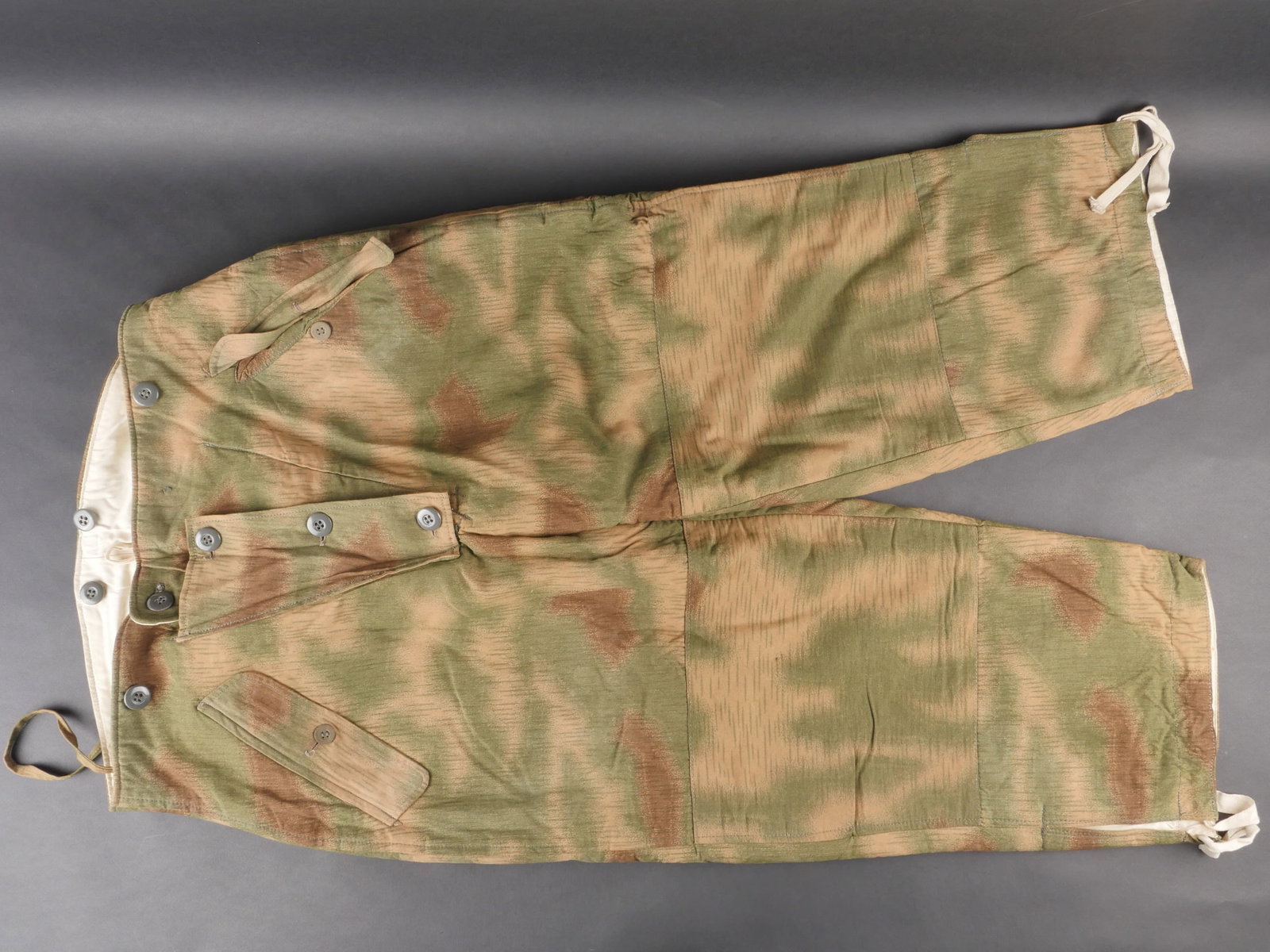 Pantalon camoufle reversible. Reversible camouflage trousers: Pantalon camoufle reversible en tissu camoufle flou avec interieur double en coton blanc. Tous les cordons et boutons sont presents. Marquage RBNr 0/1007/0155, taille 1. a noter une legere usure de la