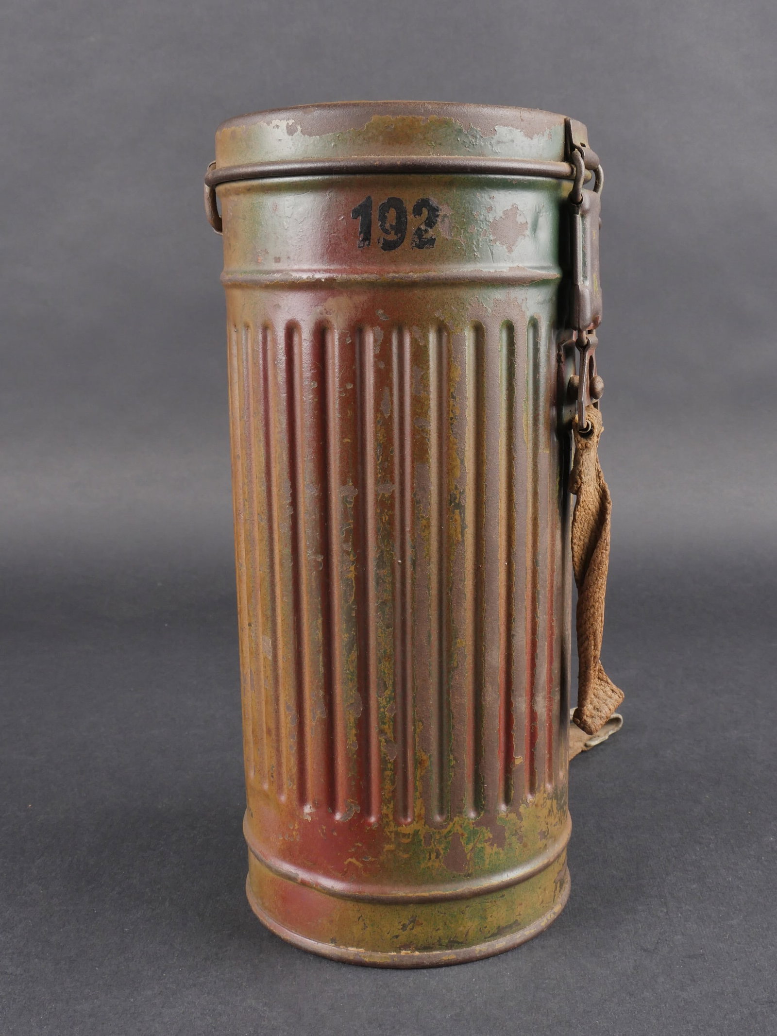 Boitier camoufle. Camouflaged gas mask box.: Boitier camoufle de masque a gaz allemand, modele long en metal. Tirette et petite sangle presentes, grande sangle manquante. Peinture camouflee trois tons vert, sable et ocre conservee a 50 %. Numero