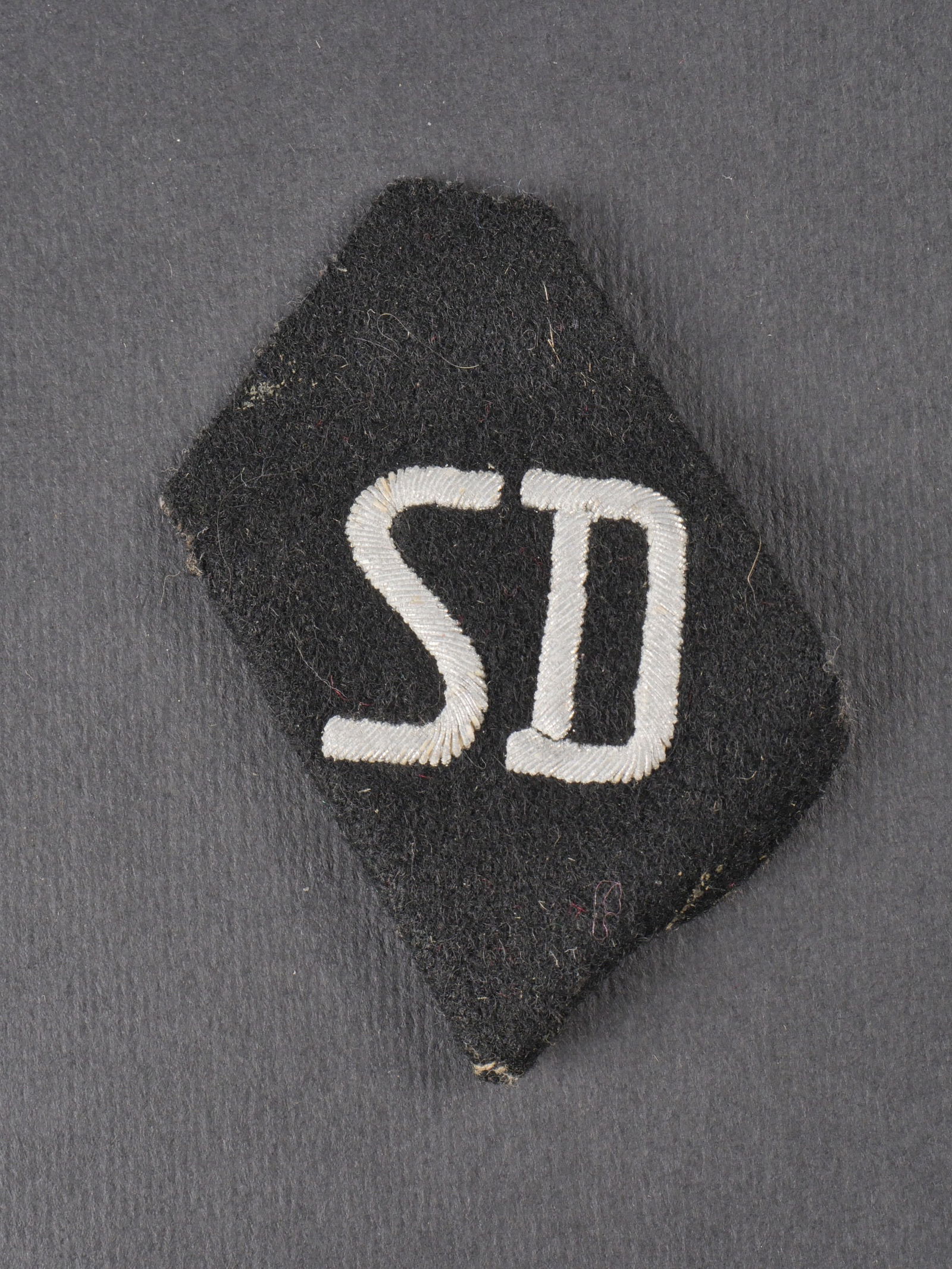 Insigne de manche SD. SD sleeve insignia.: Insigne de manche SD en losange de drap noir, lettrage SD en cannetille argent, bordures partiellement defaites. Usure et patine visibles. etat II+. SD sleeve insignia in black wool diamond, silver bu
