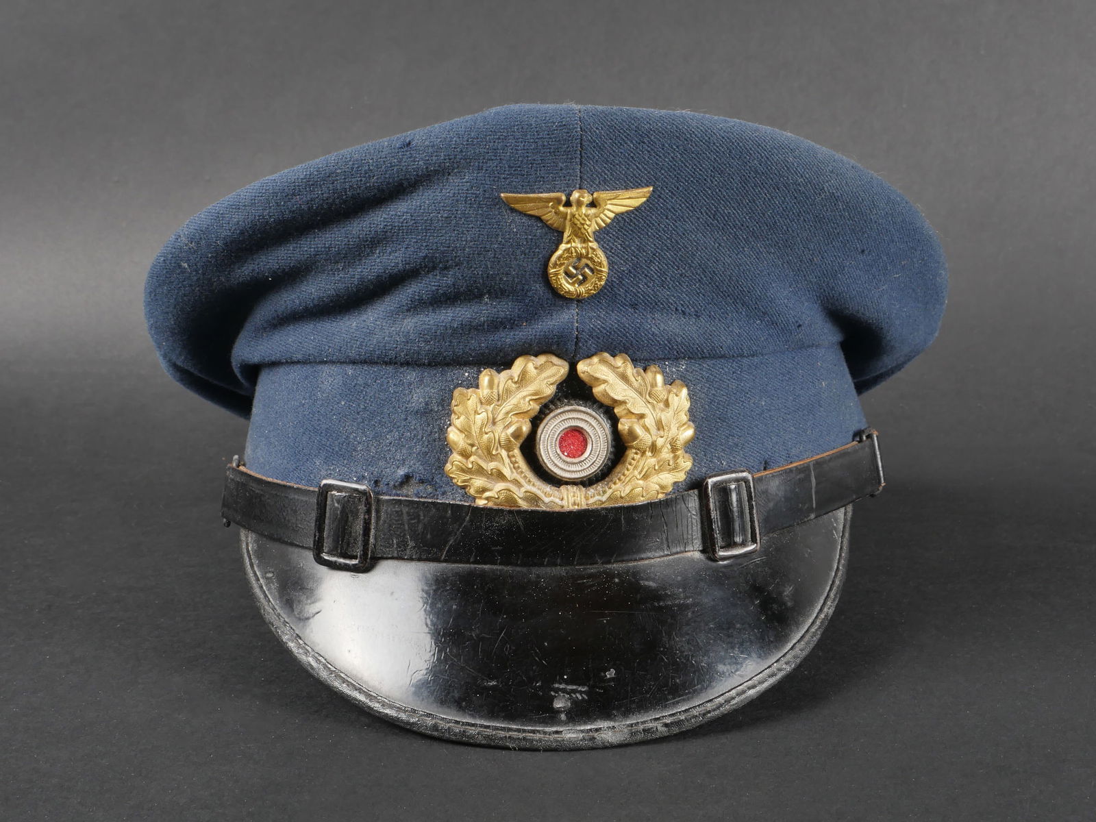 Casquette allemande. German Cap.: Casquette allemande en drap bleu ciel, ornee d’insignes metalliques dores dont les fixations n’ont pas ete demontees. Fausse jugulaire en cuir noir, visiere complete. Bandeau de sudation en simili