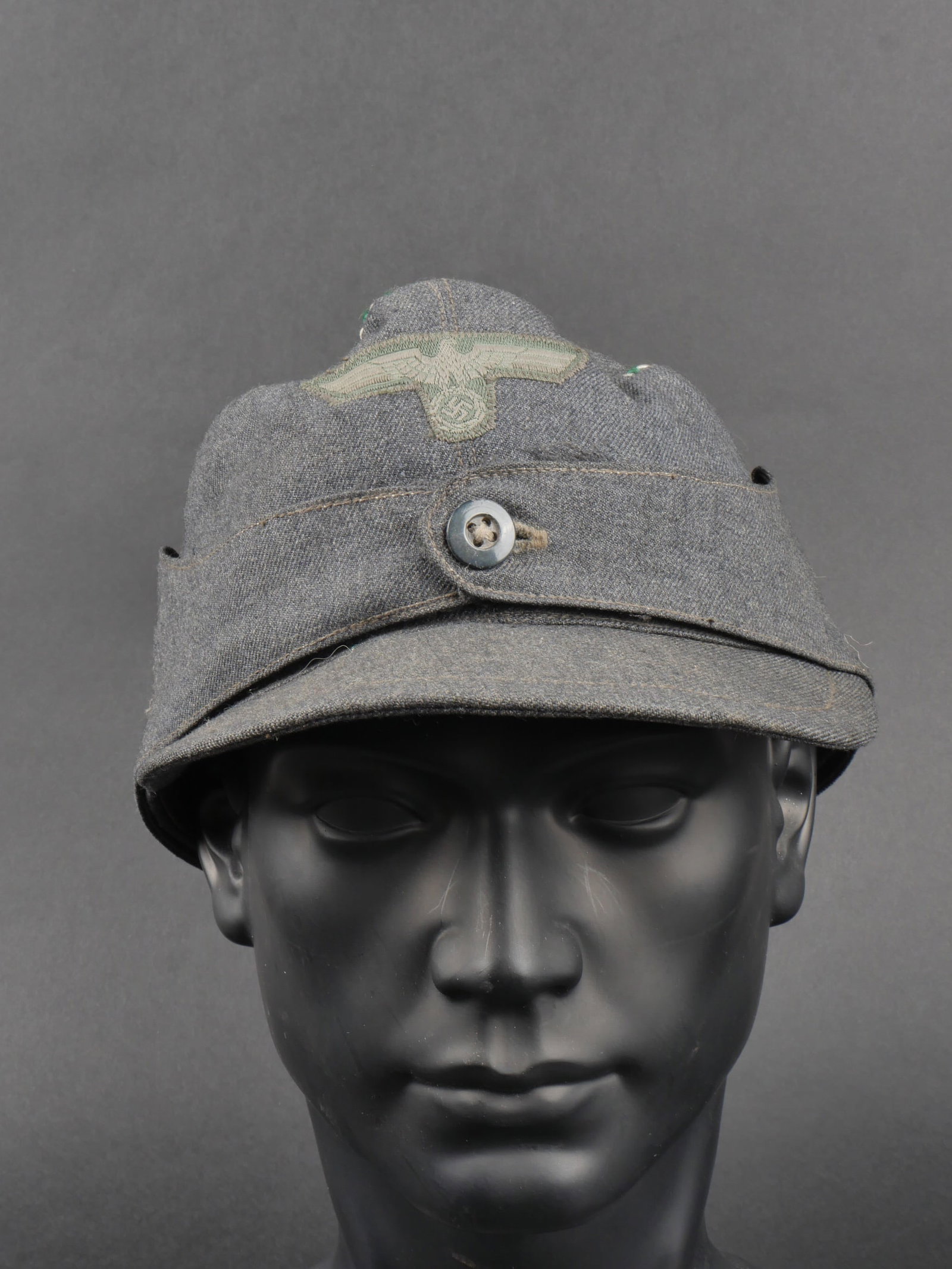 Casquette allemande. German Cap.: Casquette allemande en fin tissu de coton gris, lisere vert et argent. Aigle de la Heer cousu a la machine. Presence d’un bouton de fermeture des rabats. Visiere avec pliures, bandeau de sudation en