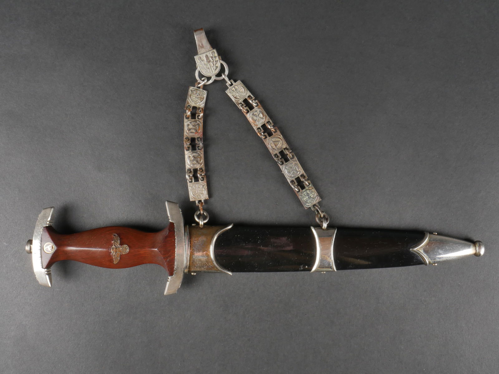 Dague NSKK a chainette. NSKK chained dagger: Dague NSKK a chainette avec garnitures en metal nickele. Poignee en bois marron surmontee d’un aigle allemand remonte et d’un insigne SA d’origine. Lame en tres bon etat, marquee Alles Fur Deuts