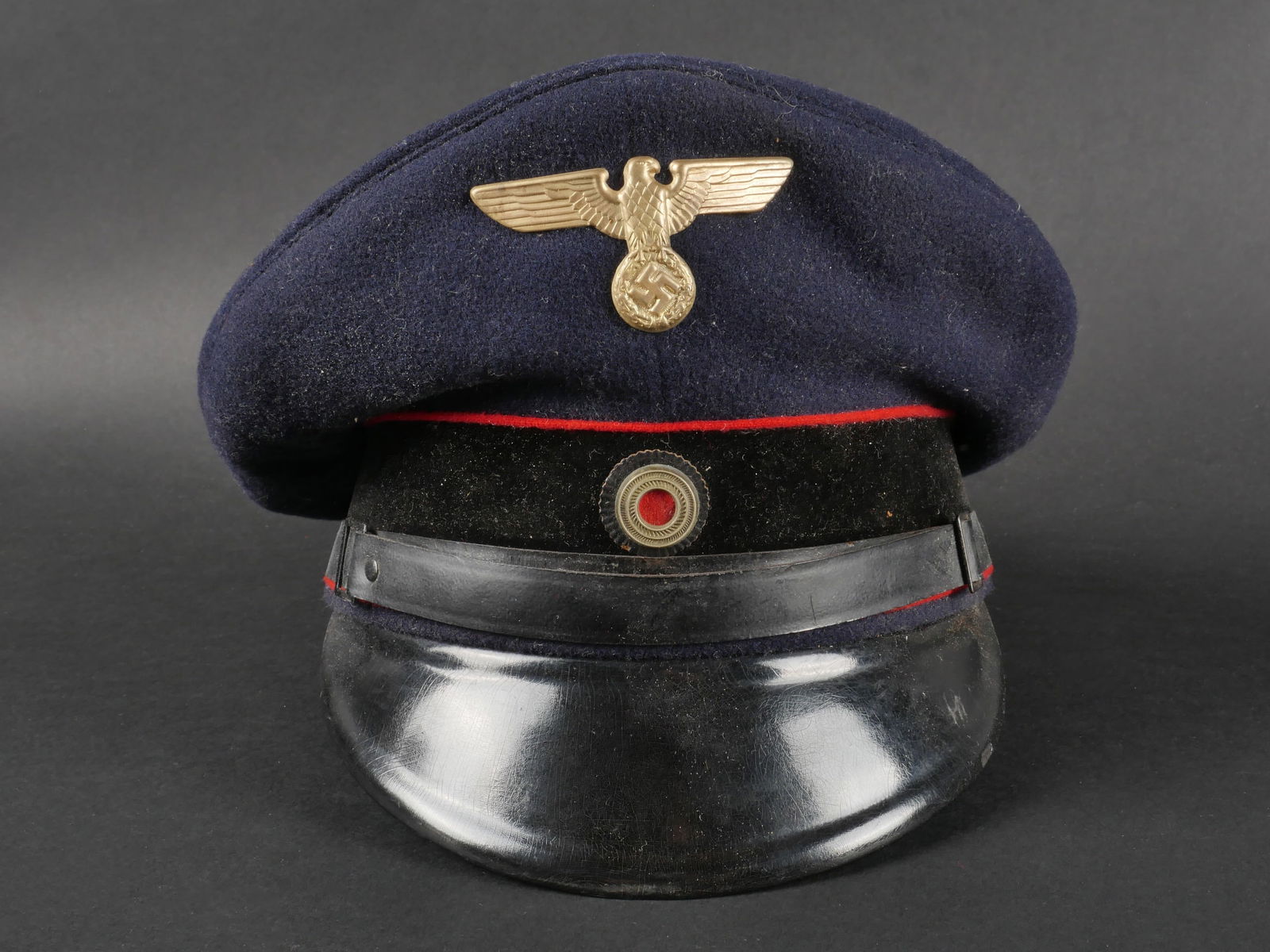 Casquette de la Reichsbahn. Reichsbahn cap: Casquette de la Reichsbahn en drap bleu marine. La partie superieure de la coiffe presente une importante reparation. Bandeau en velours noir. Insignes en metal non demontes, etat des attaches non ver