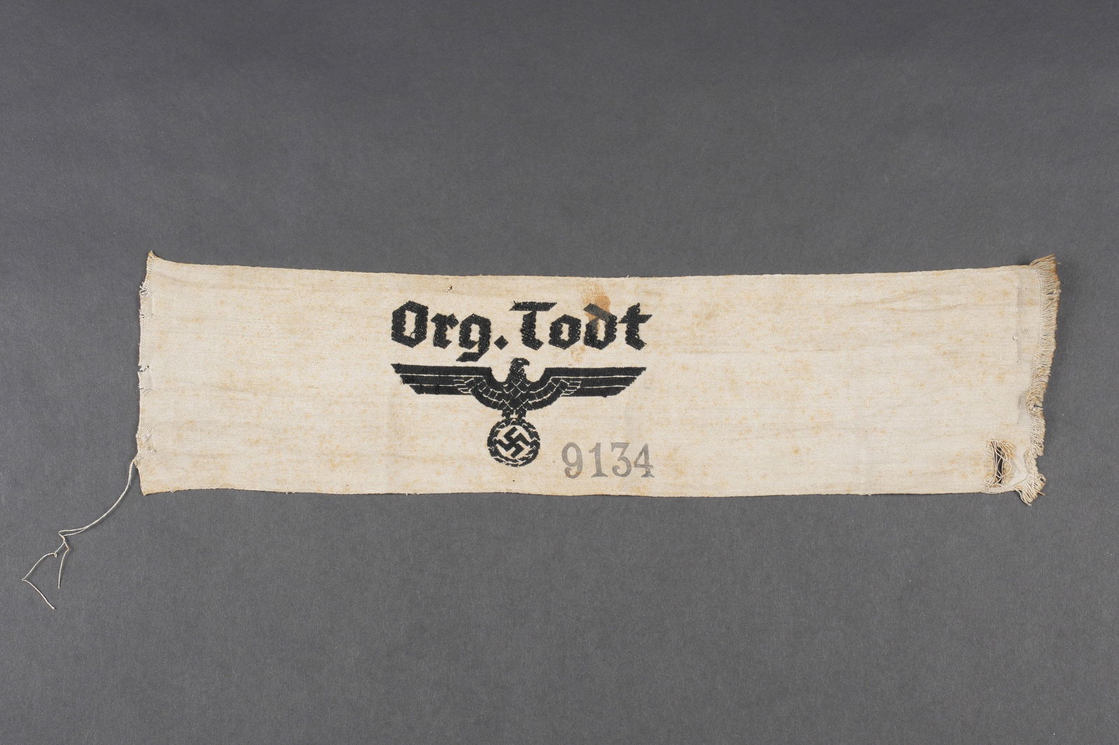 Brassard Organisation Todt. Organisation Todt armband.: Brassard de l organisation Todt. En tissu coton gris. Aigle et mention Org Todt brode. Numero 9134 imprime. La couture arriere est defaite. A noter une certaine usure et patine de la piece, ainsi que