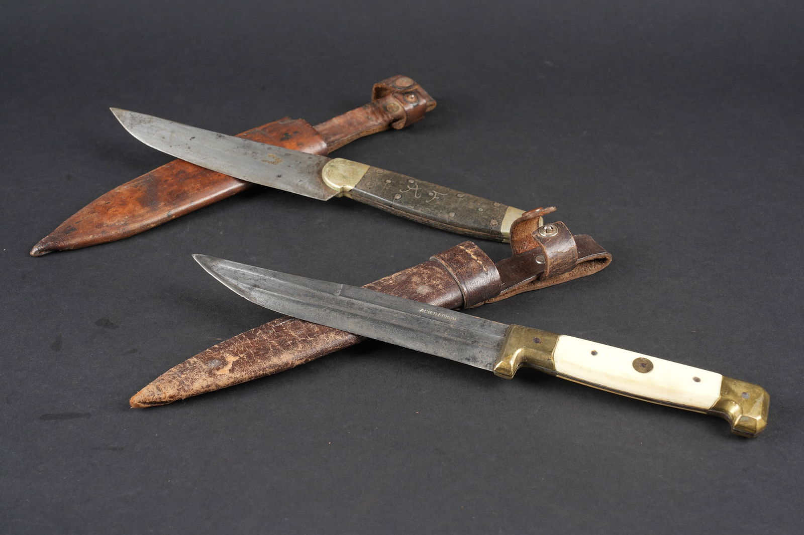 Lot de couteaux de tranchees. Lot of trench knives: Lot de couteaux de tranchees. Comprenant un couteau avec lame portant le marquage Acier fondu, manche en laiton avec plaquettes en corne. Fourreau en cuir de fabrication posterieure avec une pression
