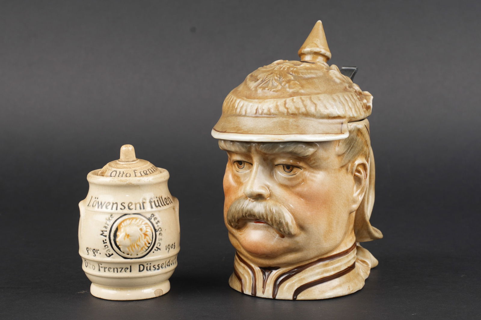 Chope a l effigie de Otto von Bismarck et pot a moutarde. Tankard bearing the likeness of Otto von: Chope anthropomorphe a l effigie de Otto von Bismarck. Chope en faience figurant Bismarck en tenue et casque de Kurassier. Le couvercle en forme de casque s ouvre avec une piece de pouce en metal fonc
